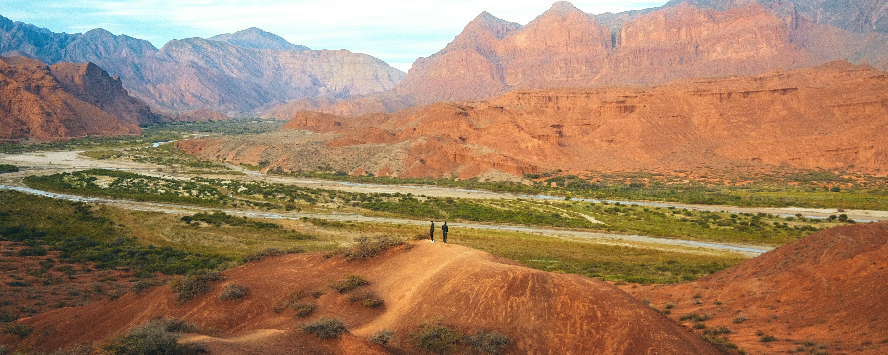 Wide valley with red hills and jagged mountains stretches beneath a pale blue sky.