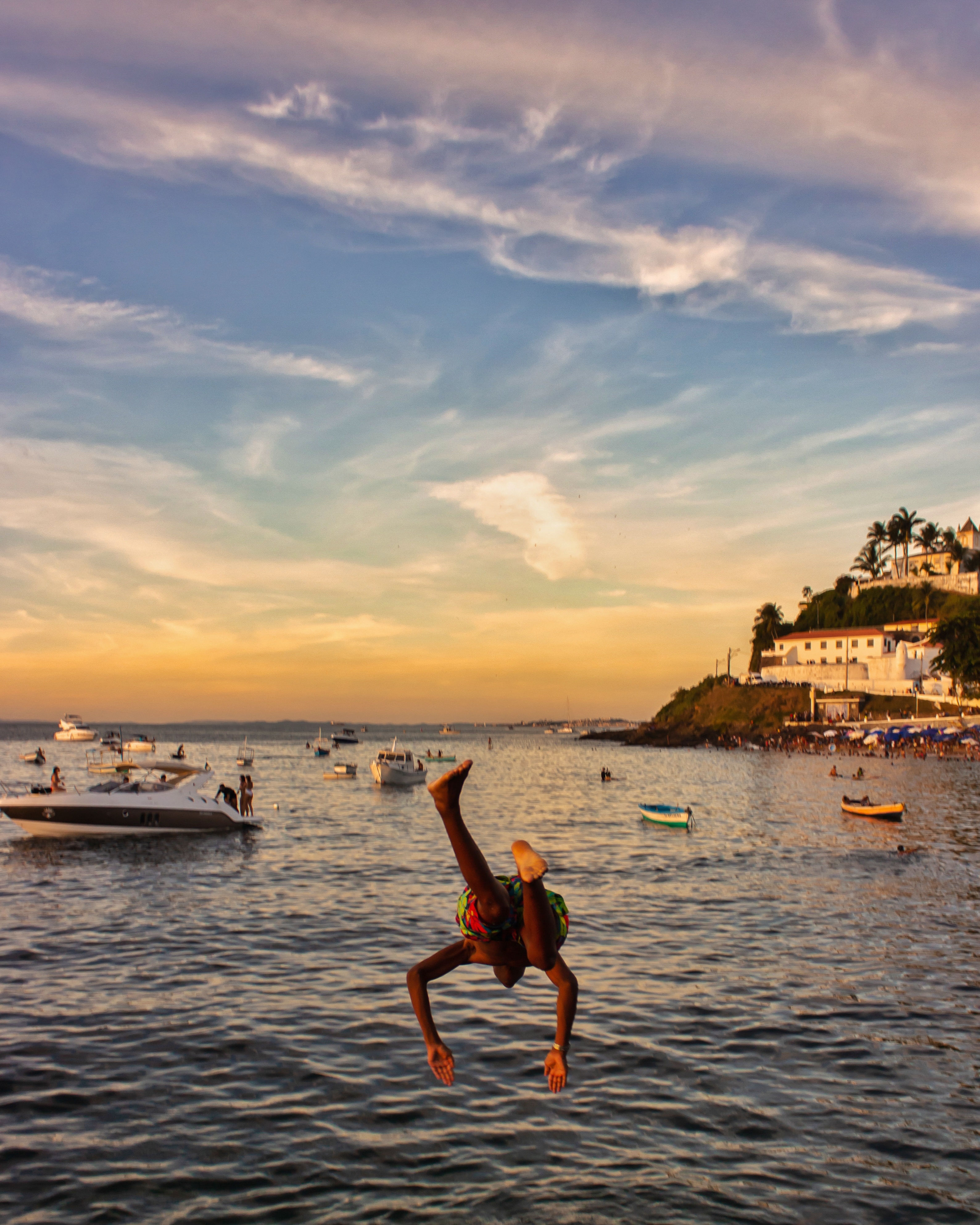 Person diving in water at sunset with boats and a shoreline in the distance.
