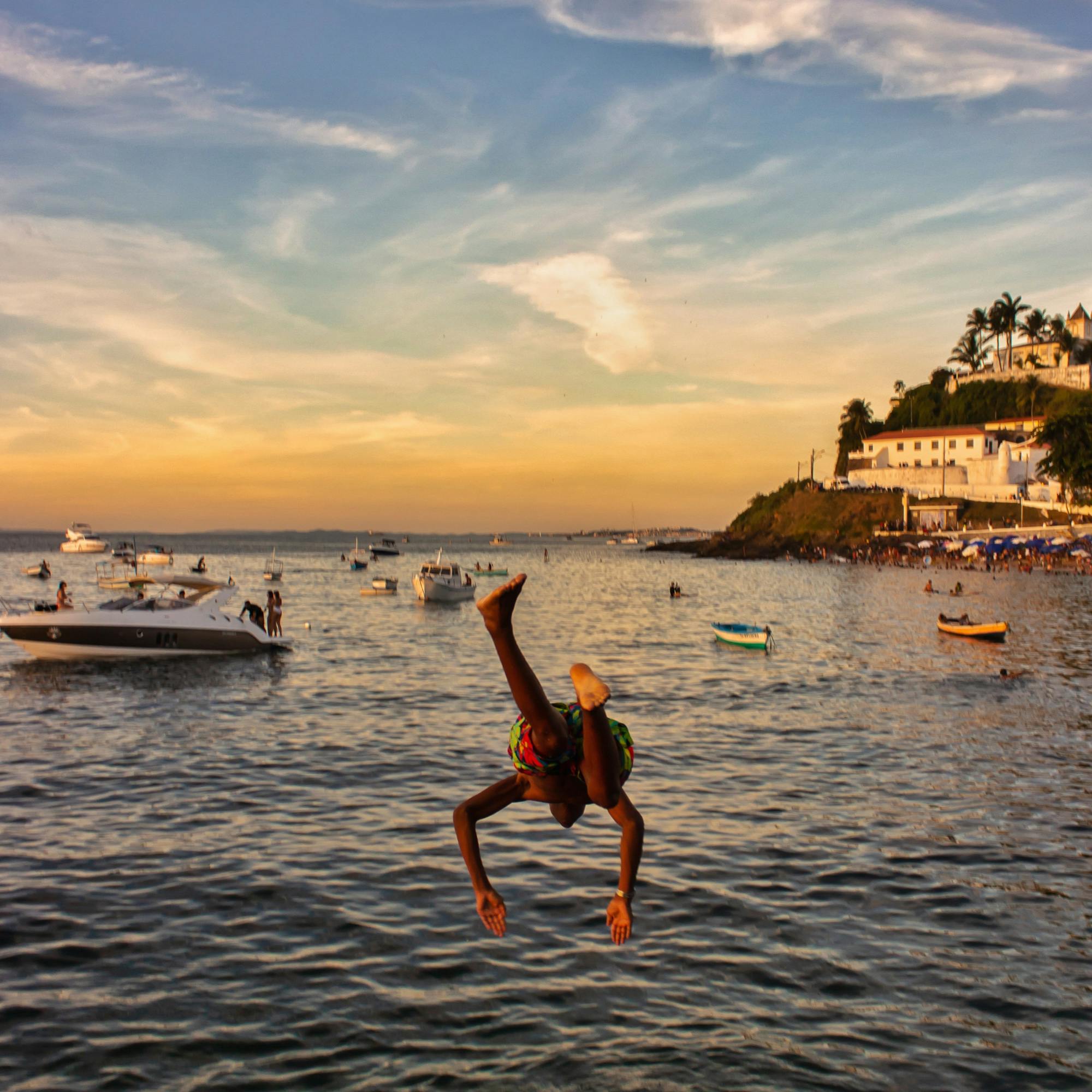 Person diving in water at sunset with boats and a shoreline in the distance.