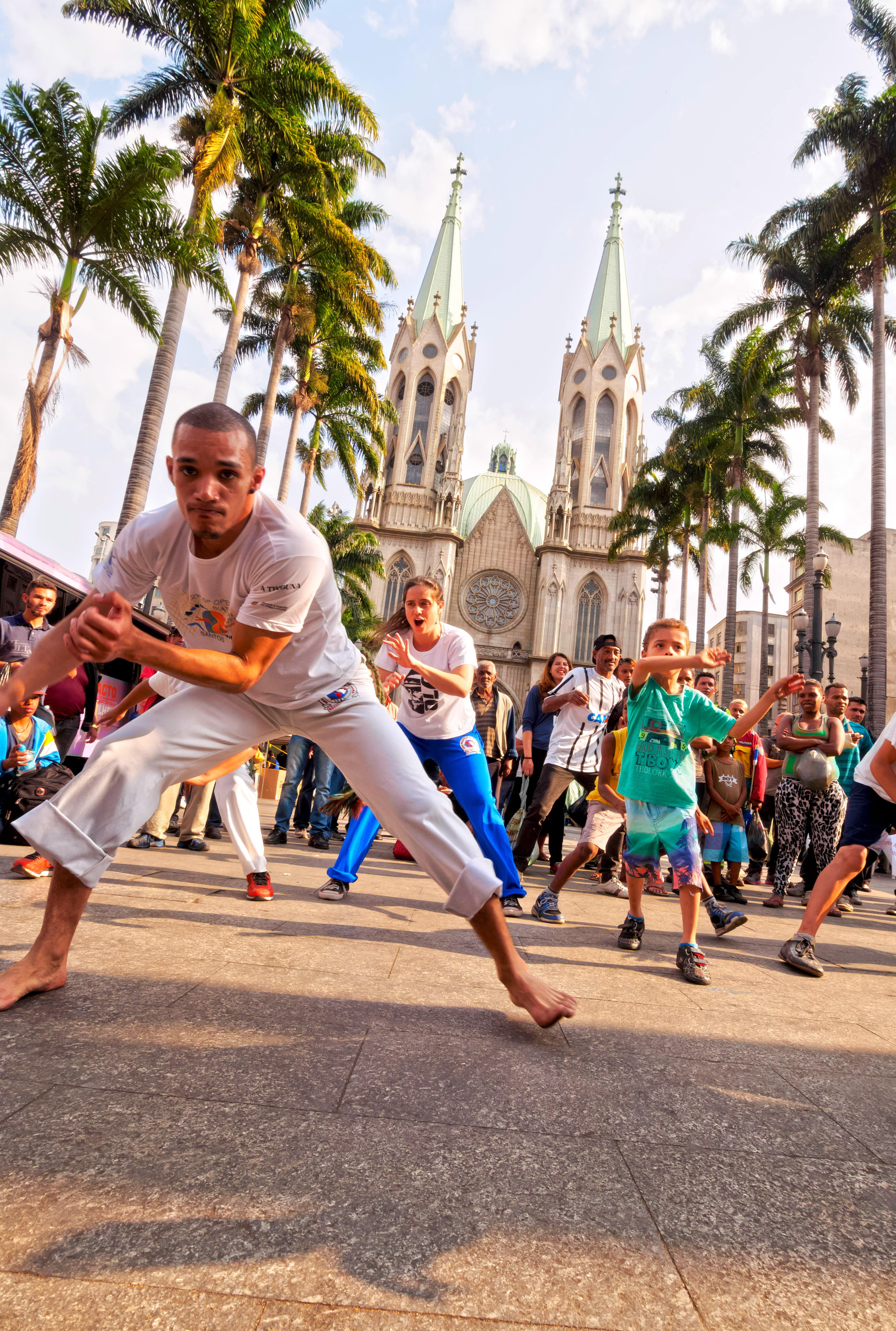 People practice capoeira on a street as others watch, with a church tower in the background.
