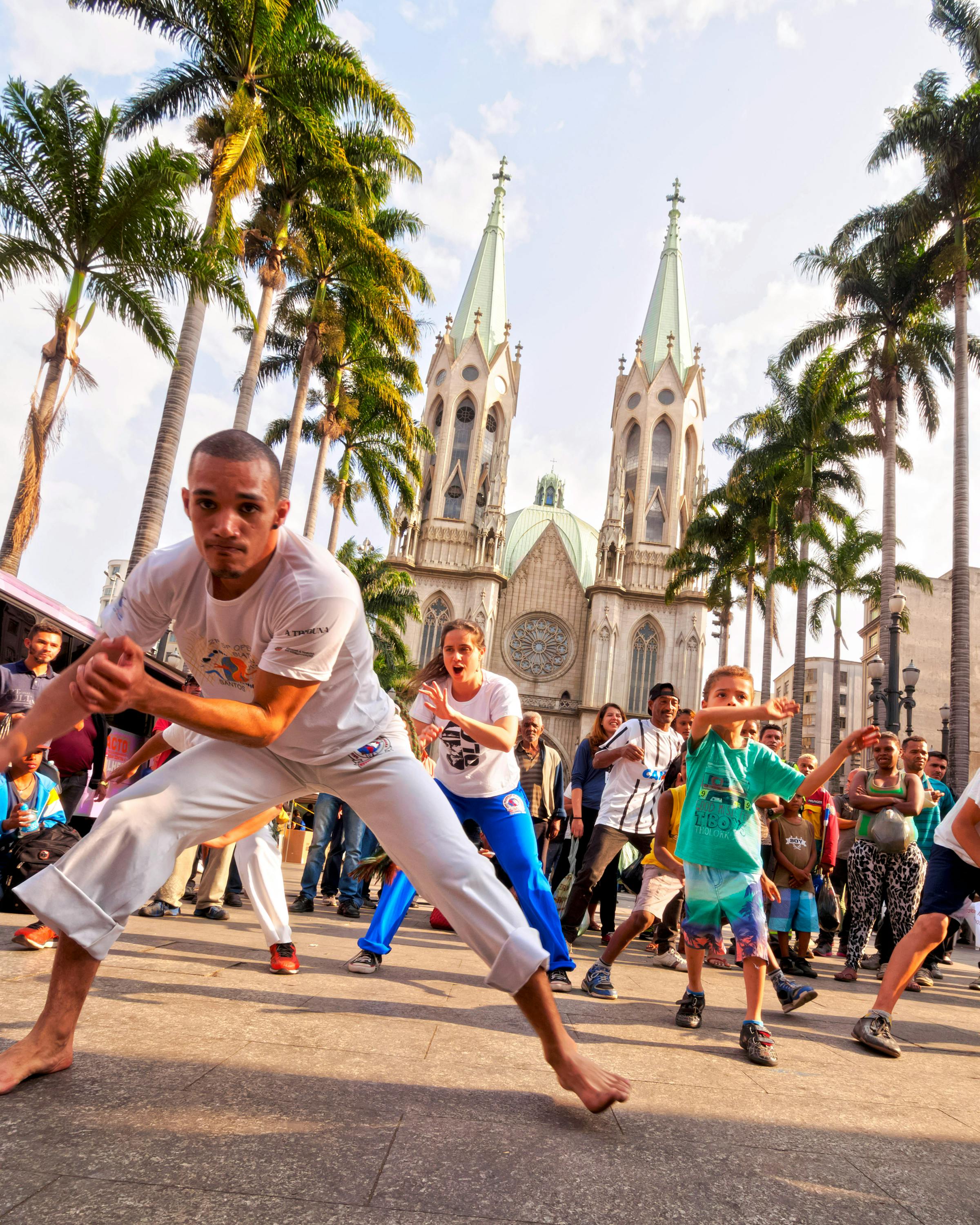 People practice capoeira on a street as others watch, with a church tower in the background.