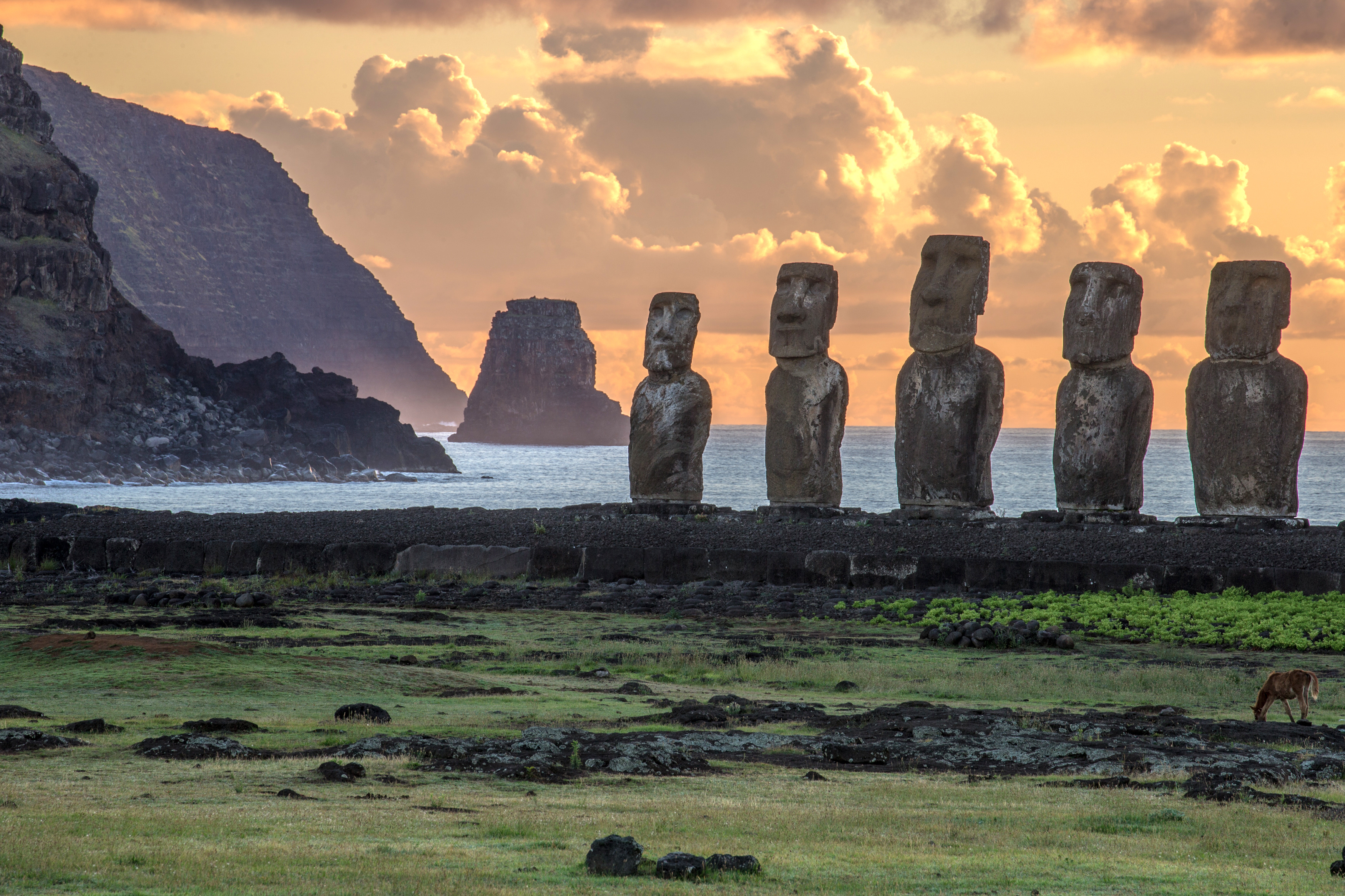 Stone statues stand on grassy ground near the ocean at sunset with clouds glowing overhead.