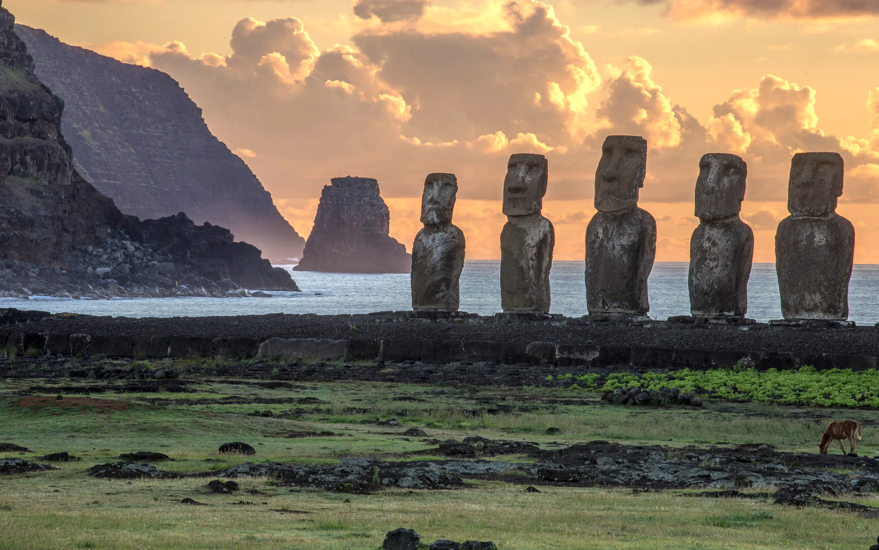 Stone statues stand on grassy ground near the ocean at sunset with clouds glowing overhead.