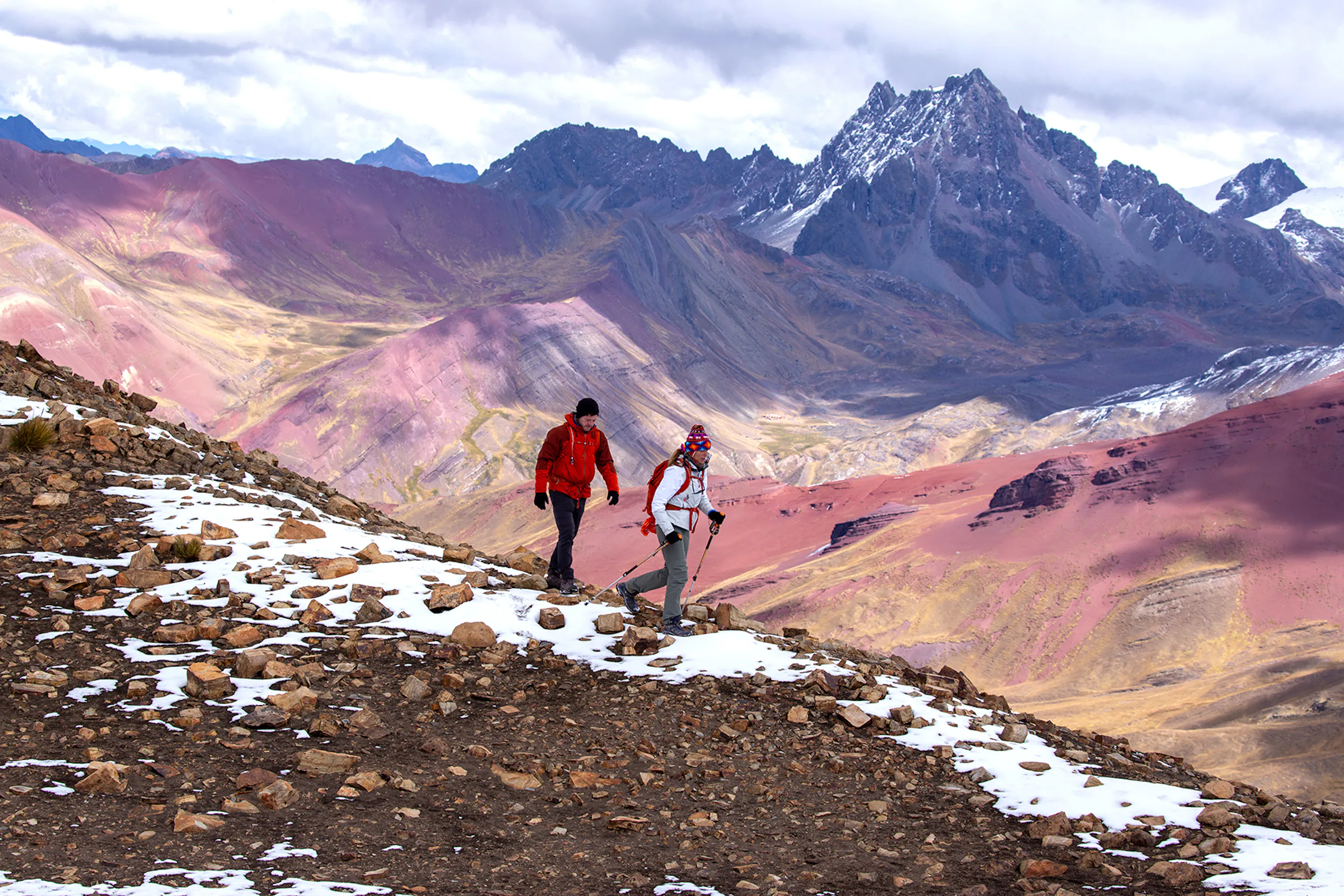 Two hikers walk along a rocky trail with snow patches and colorful mountains in the distance.