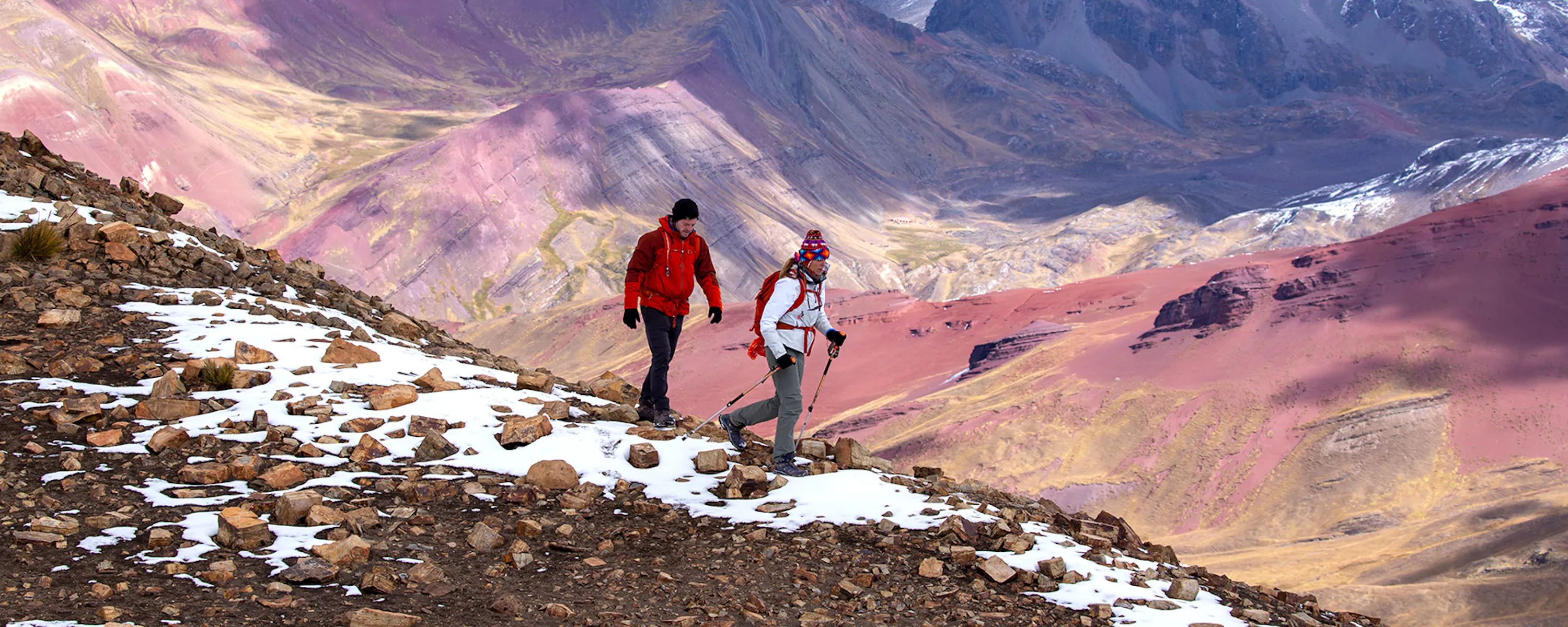 Two hikers walk along a rocky trail with snow patches and colorful mountains in the distance.