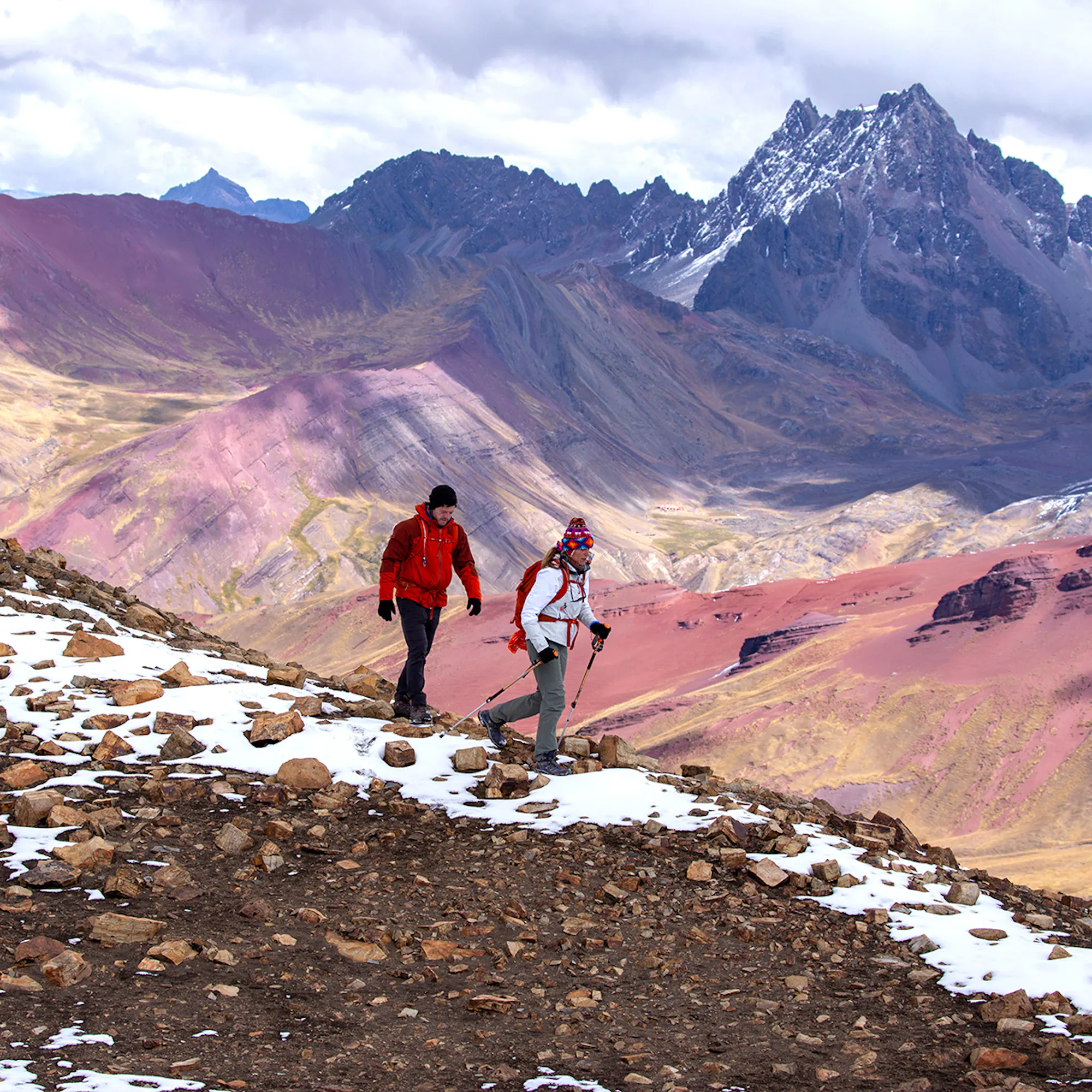 Two hikers walk along a rocky trail with snow patches and colorful mountains in the distance.