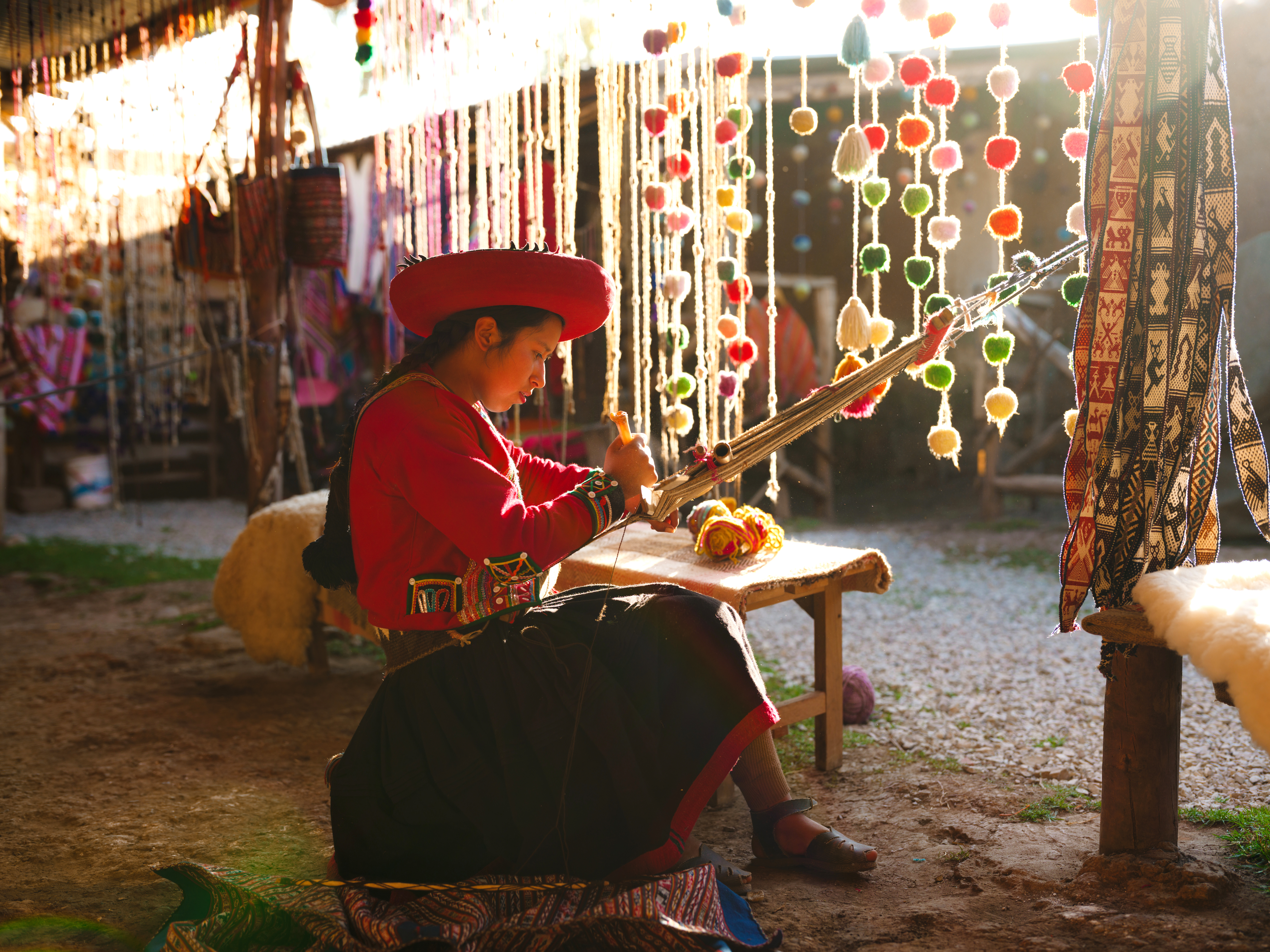 Person in traditional clothing sits weaving at a loom surrounded by colorful hanging textiles.
