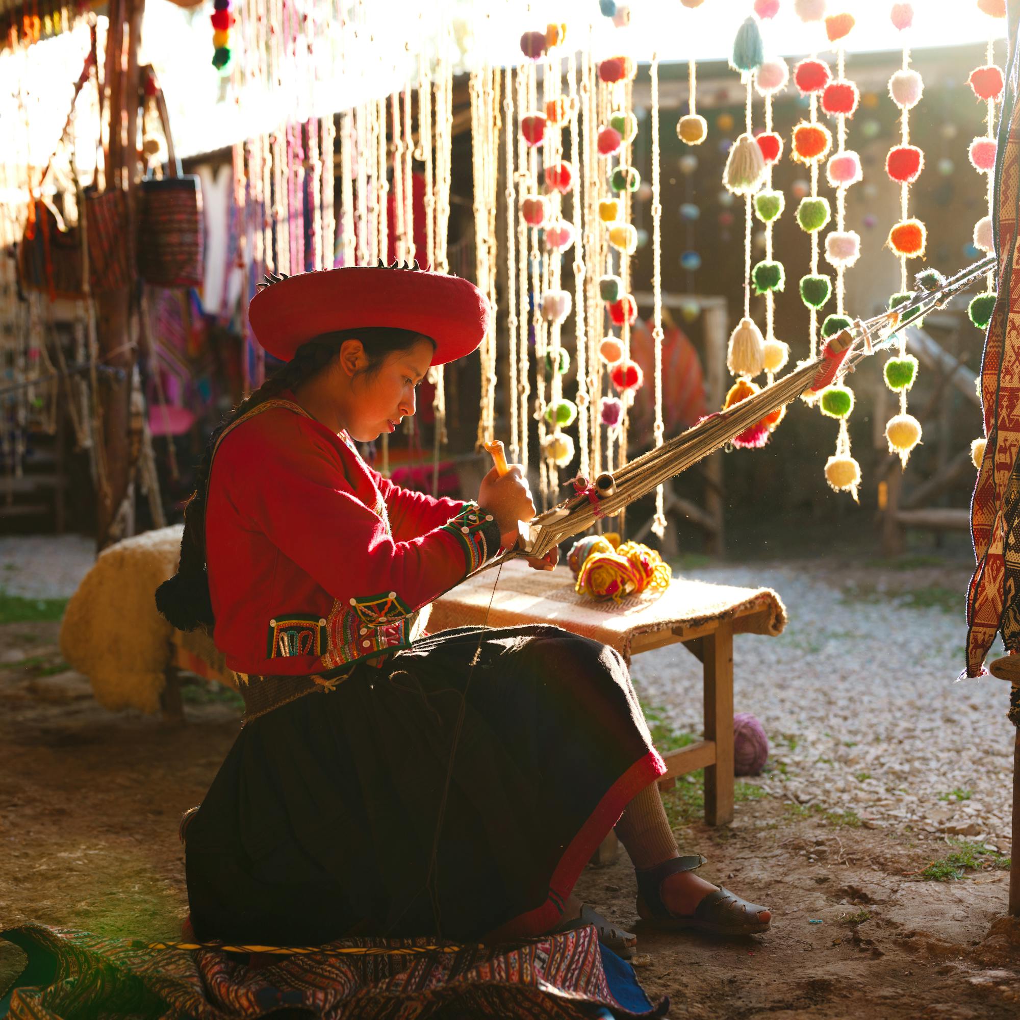 Person in traditional clothing sits weaving at a loom surrounded by colorful hanging textiles.