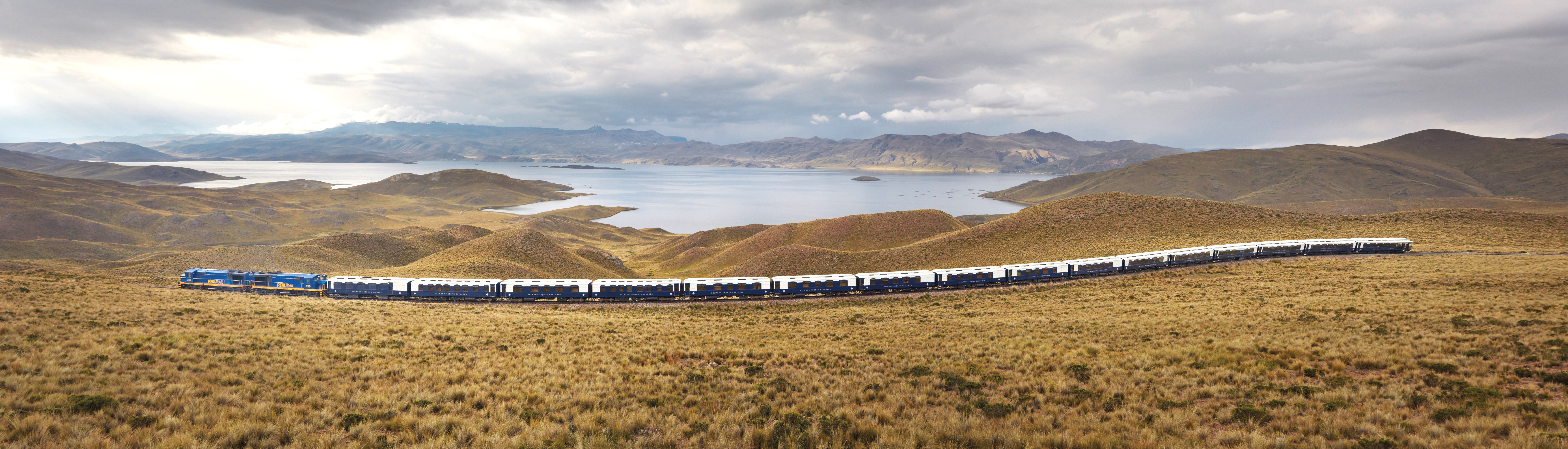 Long blue train travels across open highland plains beneath a cloudy sky.