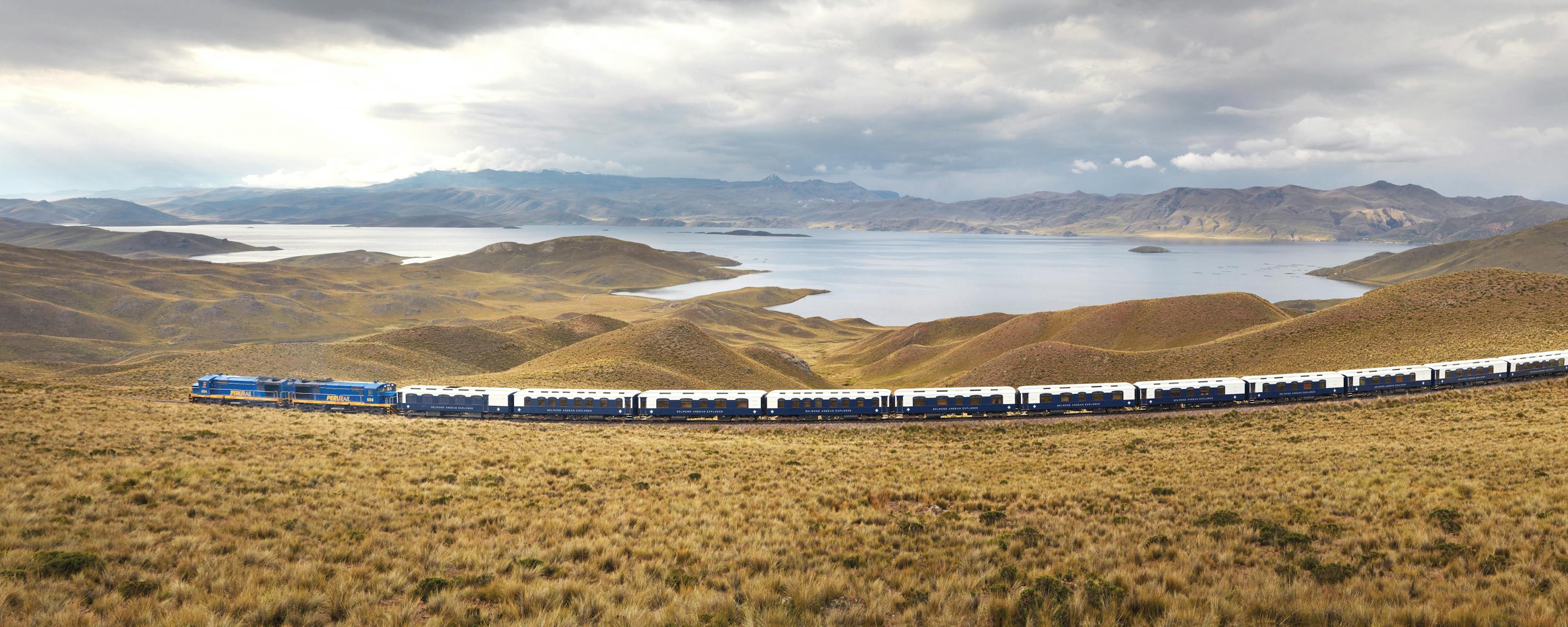 Long blue train travels across open highland plains beneath a cloudy sky.