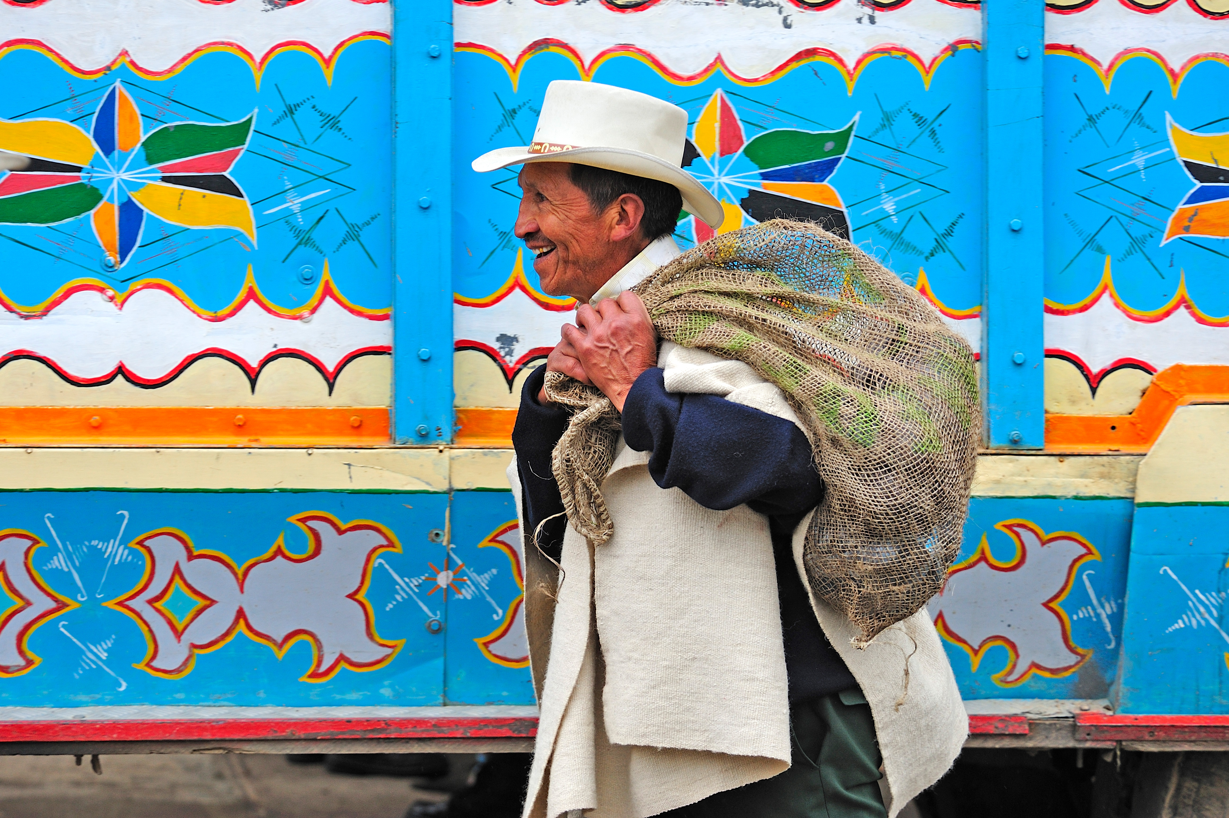 Man in a hat carries a bundle past a brightly painted wall with colorful patterns.