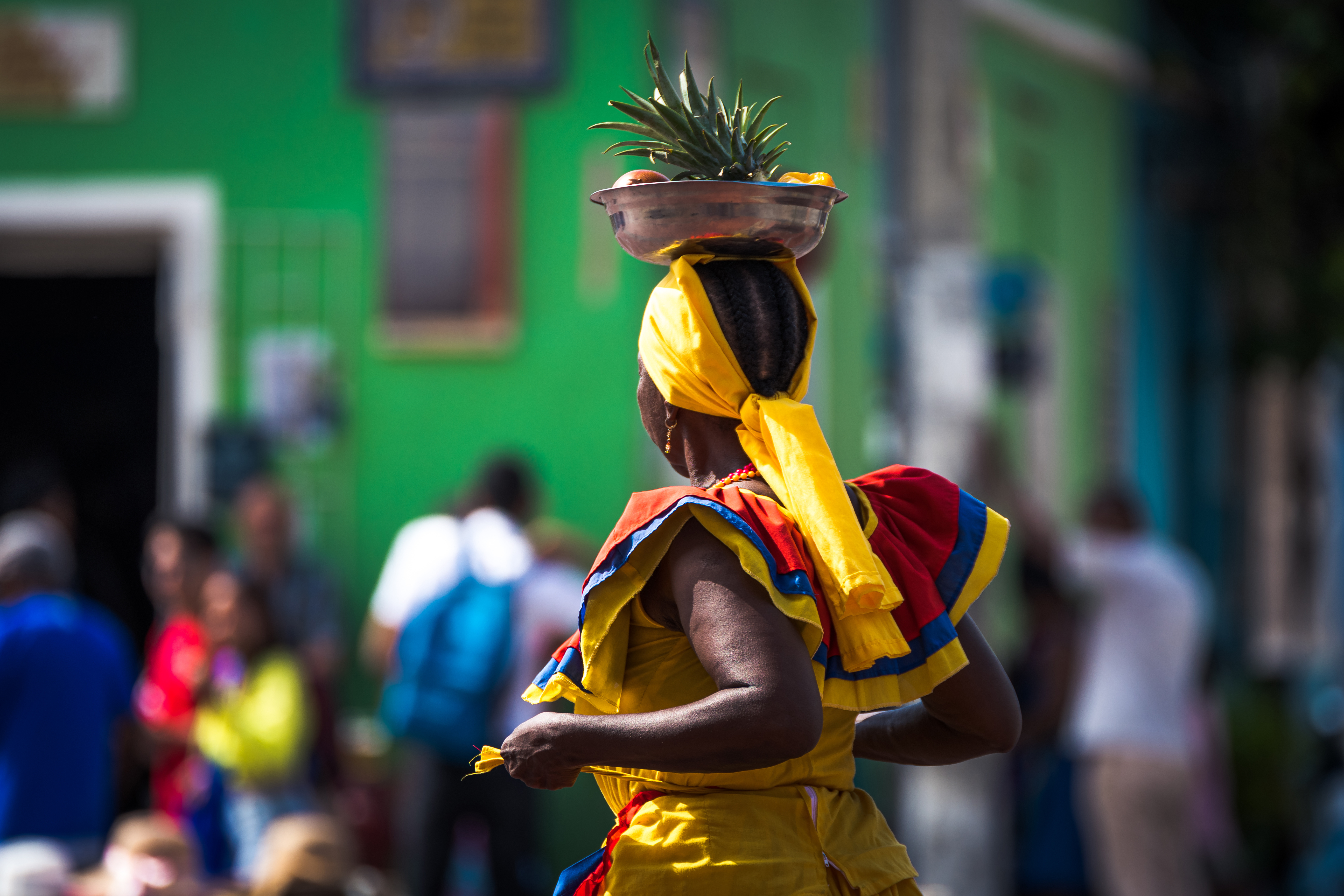 Person in bright traditional clothing carries a basket of fruit on their head in a street scene.