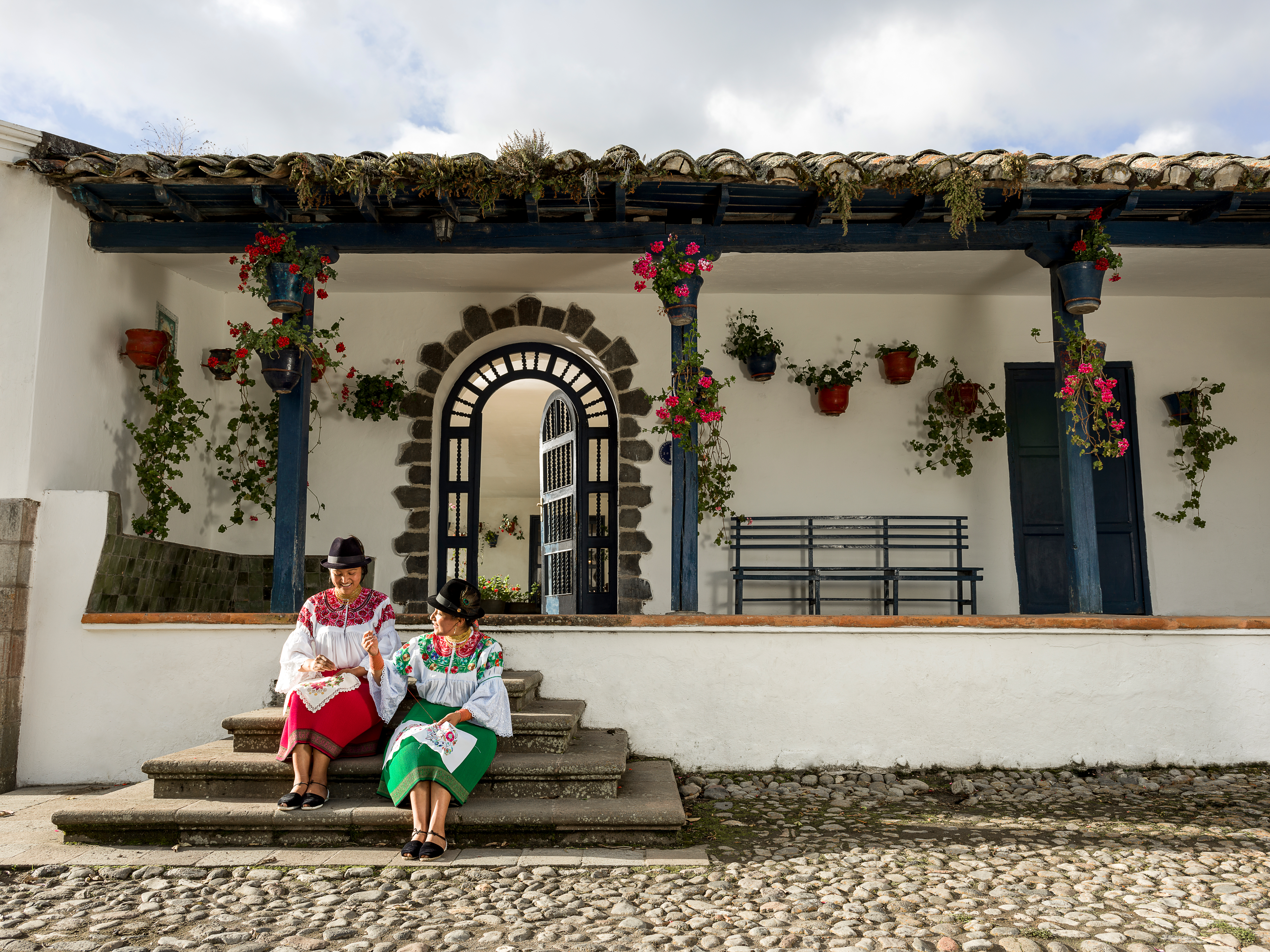 Two people sit on stone steps outside a white building with hanging flowers and an arched doorway.