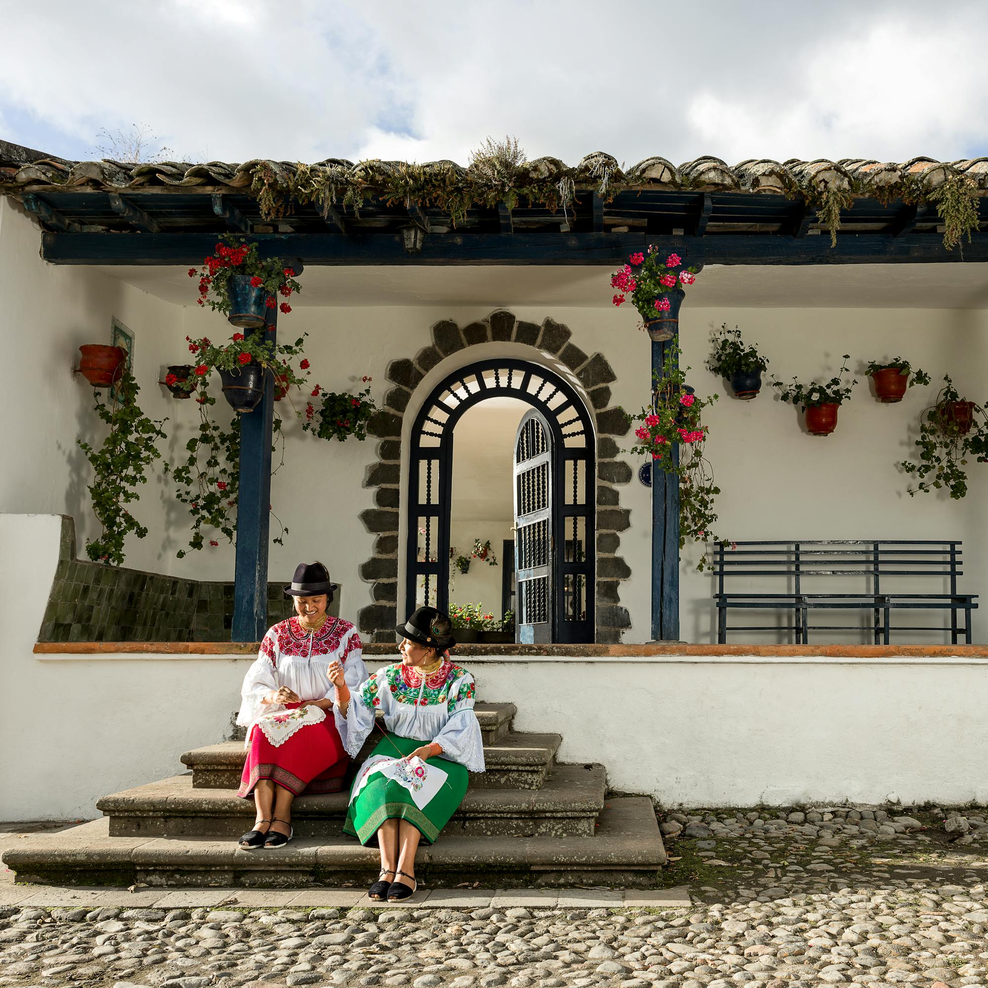 Two people sit on stone steps outside a white building with hanging flowers and an arched doorway.