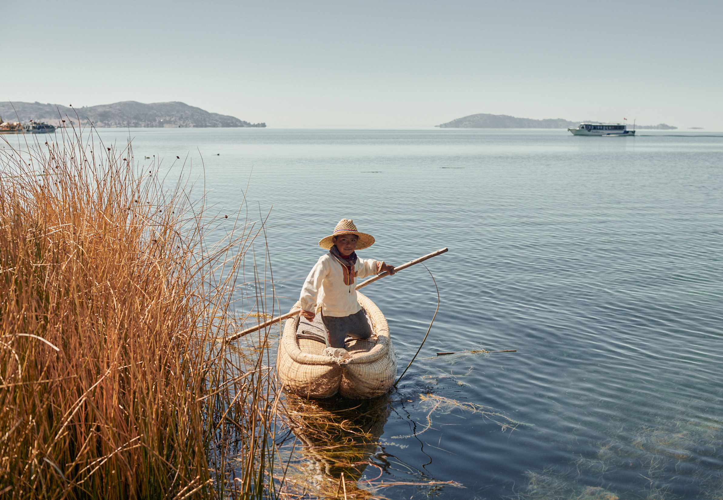 Person in a small boat glides on calm water beside tall reeds with distant hills beyond.