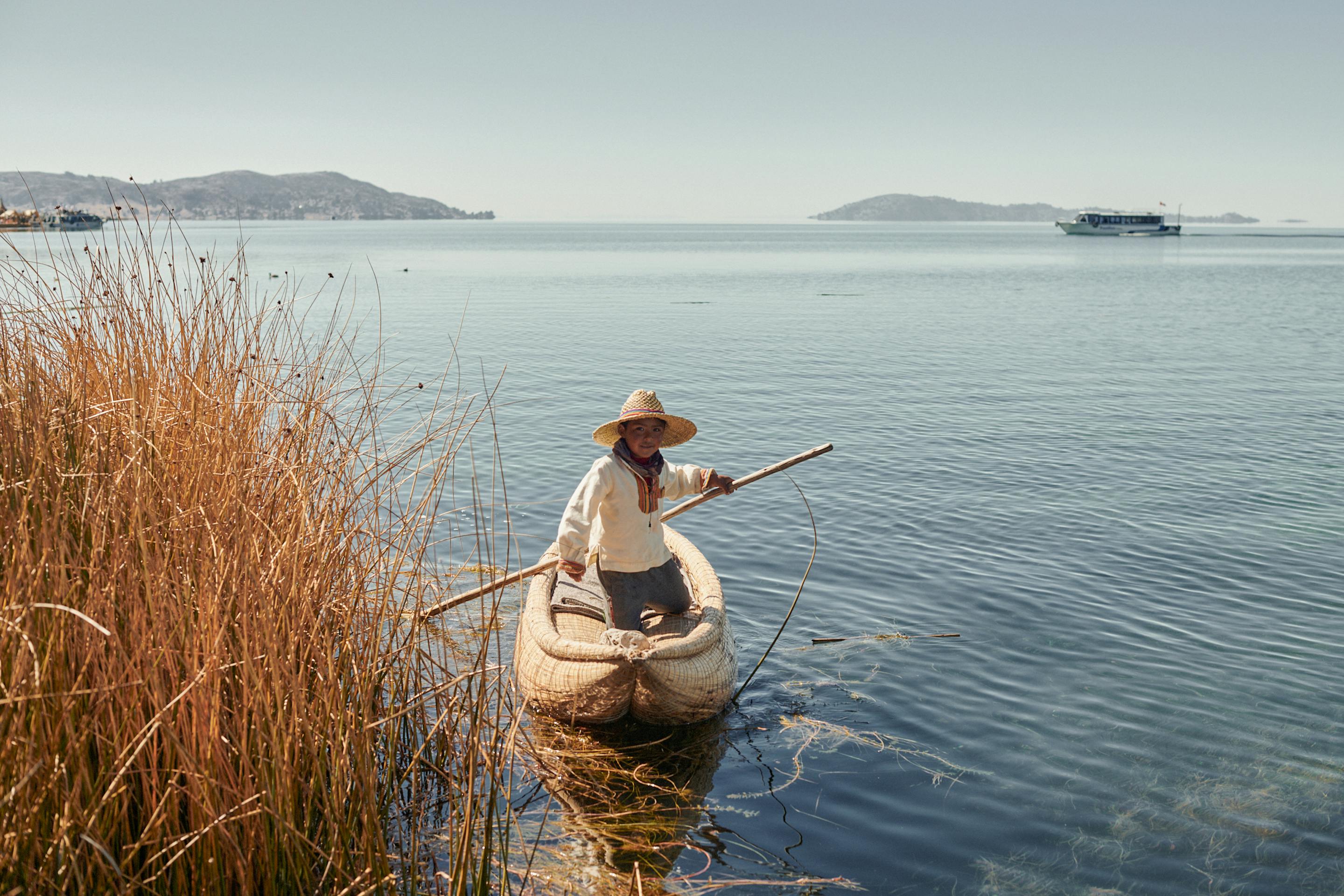 Person in a small boat glides on calm water beside tall reeds with distant hills beyond.