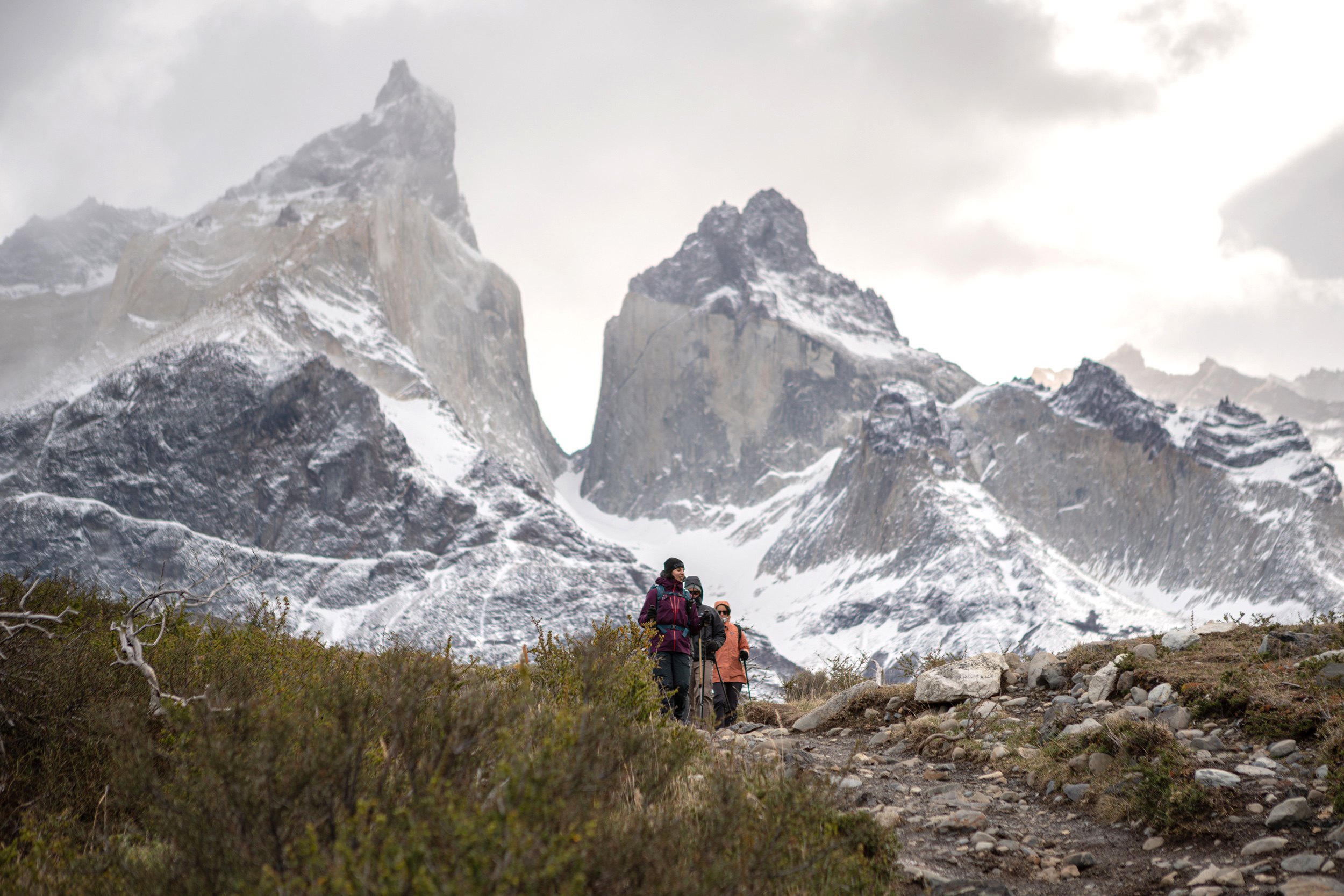 Two hikers walk up a rocky path toward jagged snow-covered peaks.