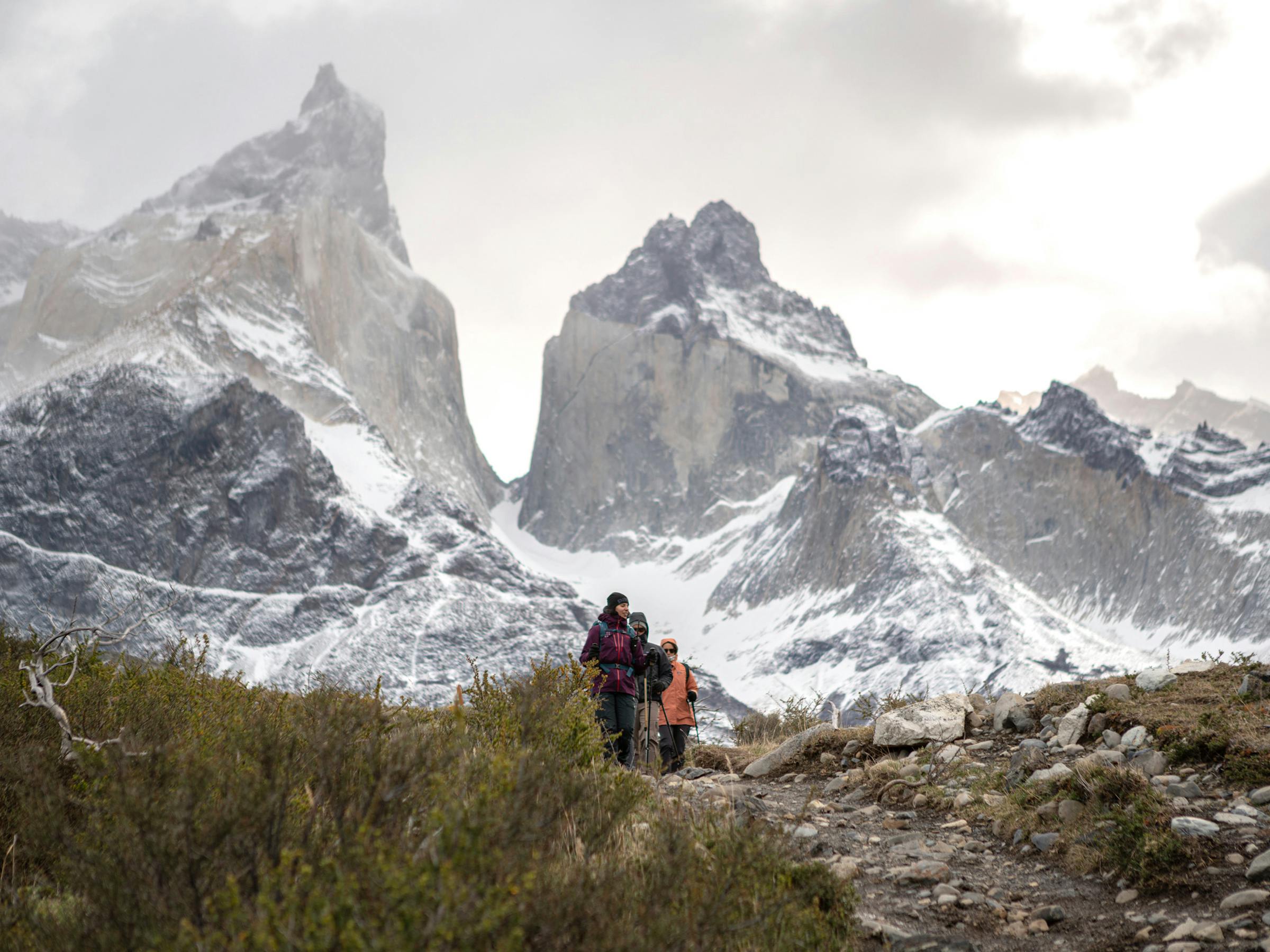 Two hikers walk up a rocky path toward jagged snow-covered peaks.
