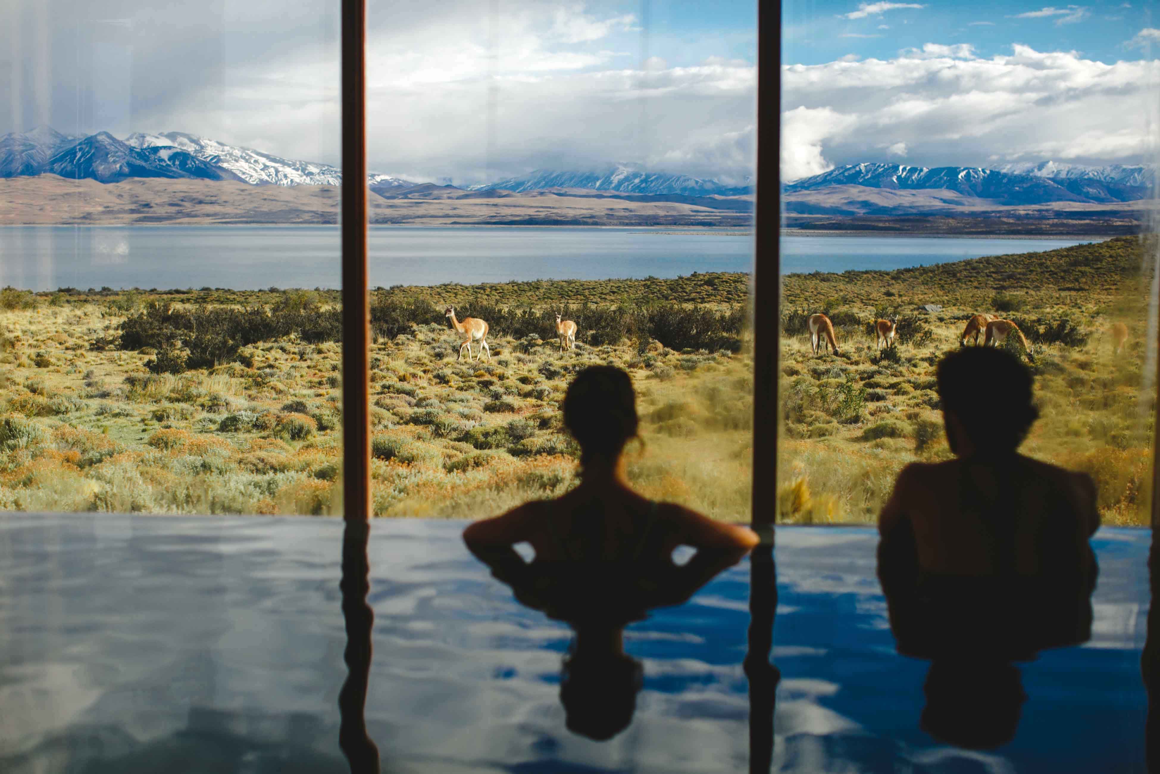 Two people relax in a pool facing floor-to-ceiling windows with a wide mountain view.