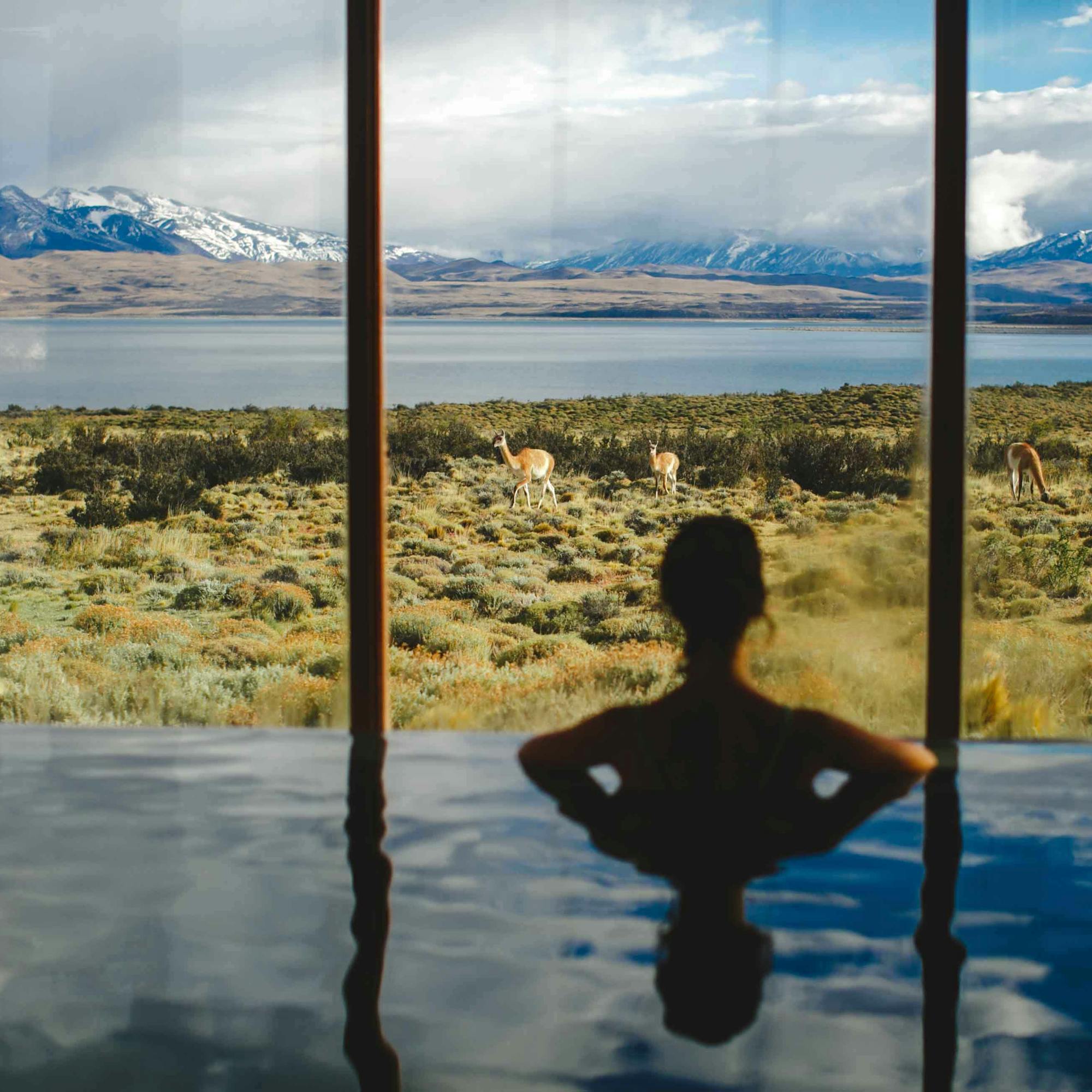 Two people relax in a pool facing floor-to-ceiling windows with a wide mountain view.