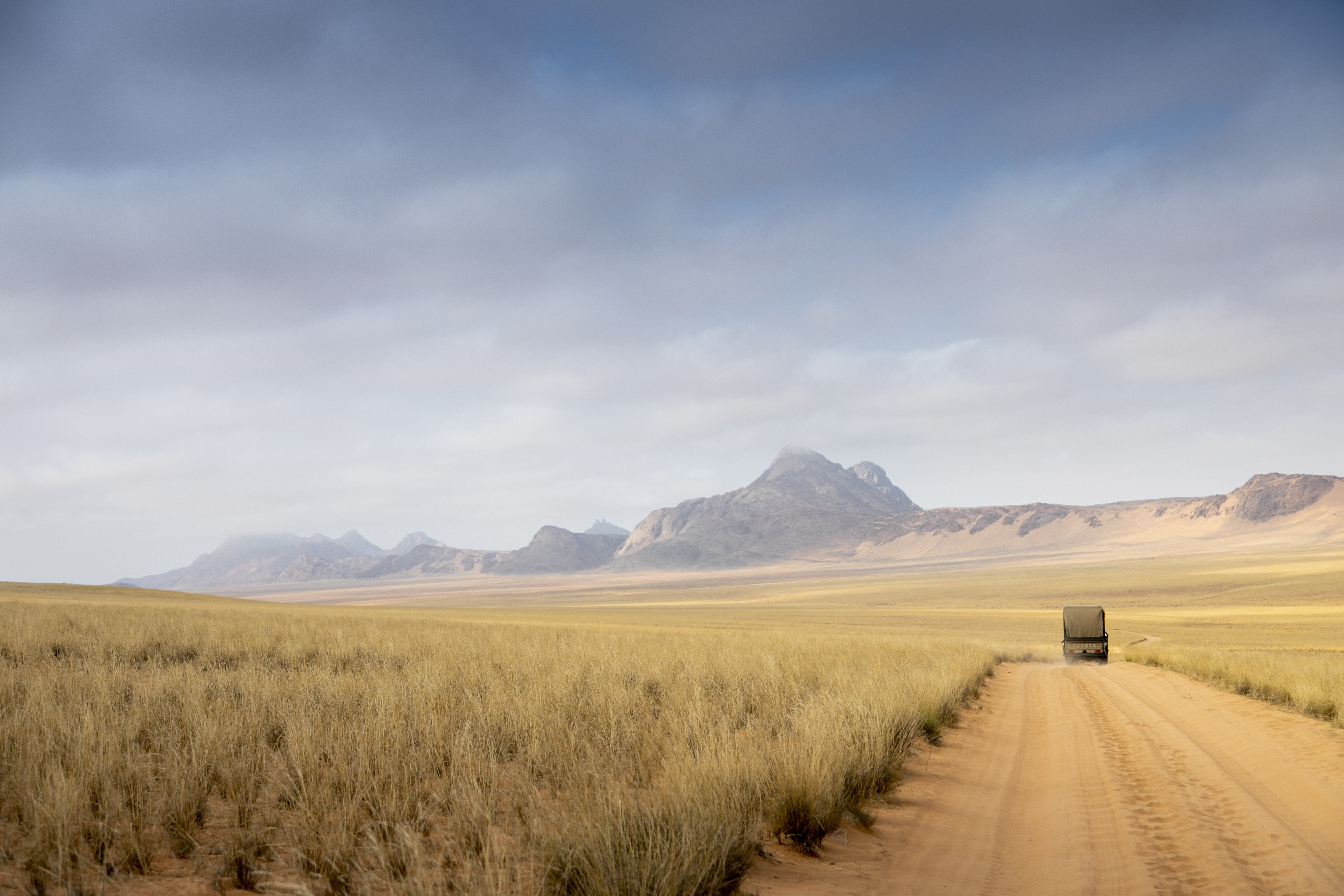 A lone vehicle travels a straight dirt road through open grassland, with blue mountains under a wide sky.