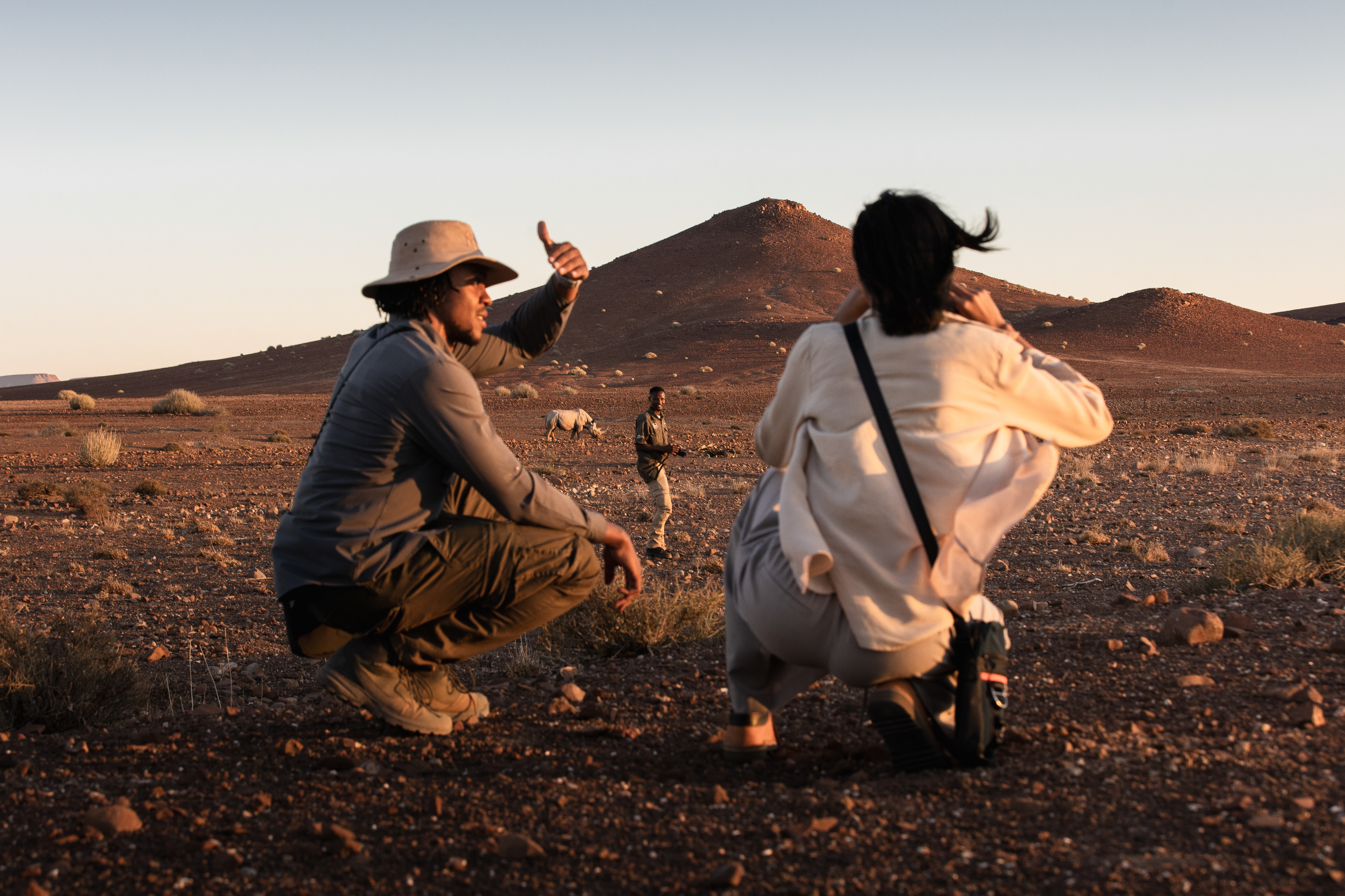 Two people crouch on dark gravel plains, pointing at tracks while red dunes and mountains glow in late light.