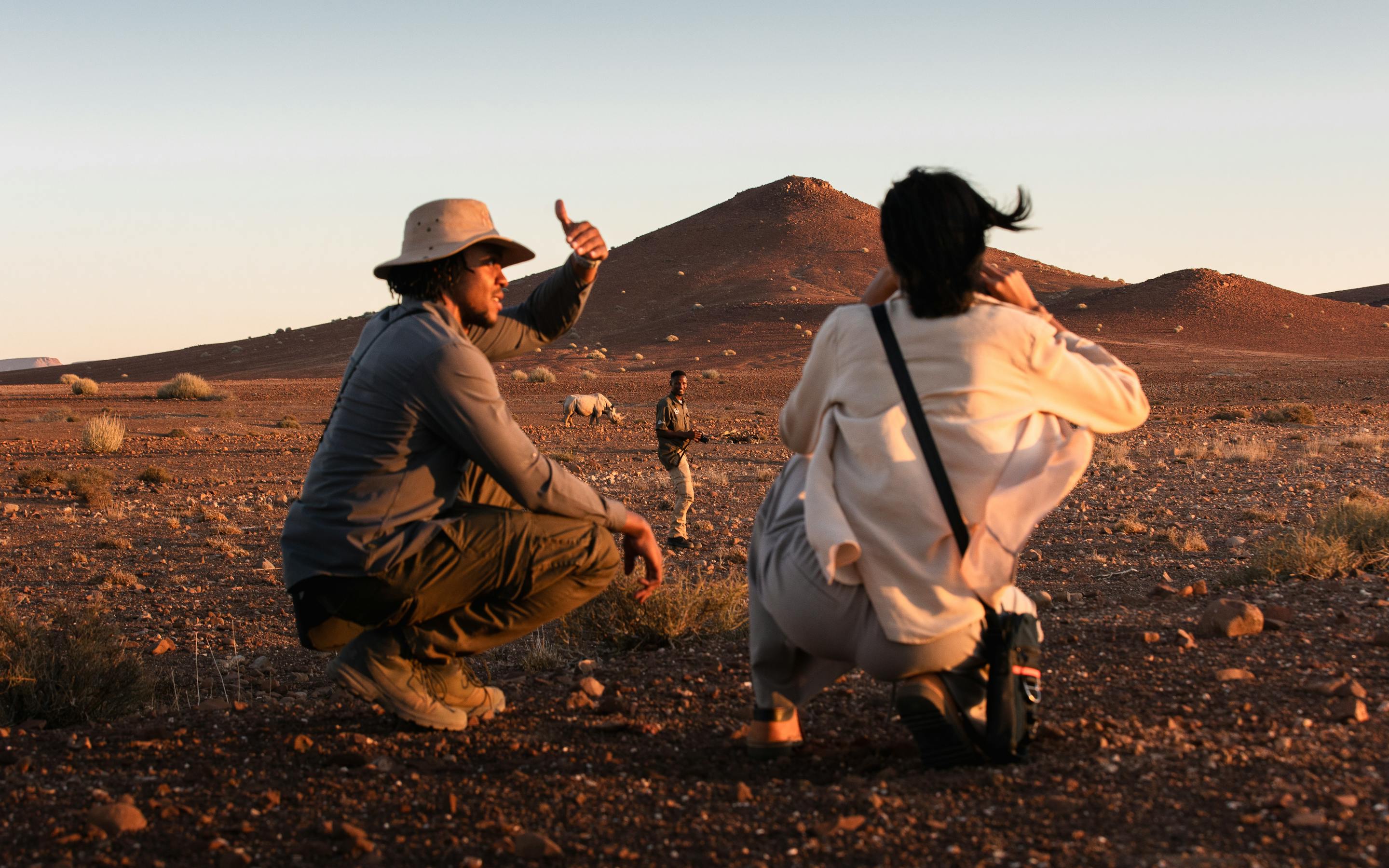 Two people crouch on dark gravel plains, pointing at tracks while red dunes and mountains glow in late light.