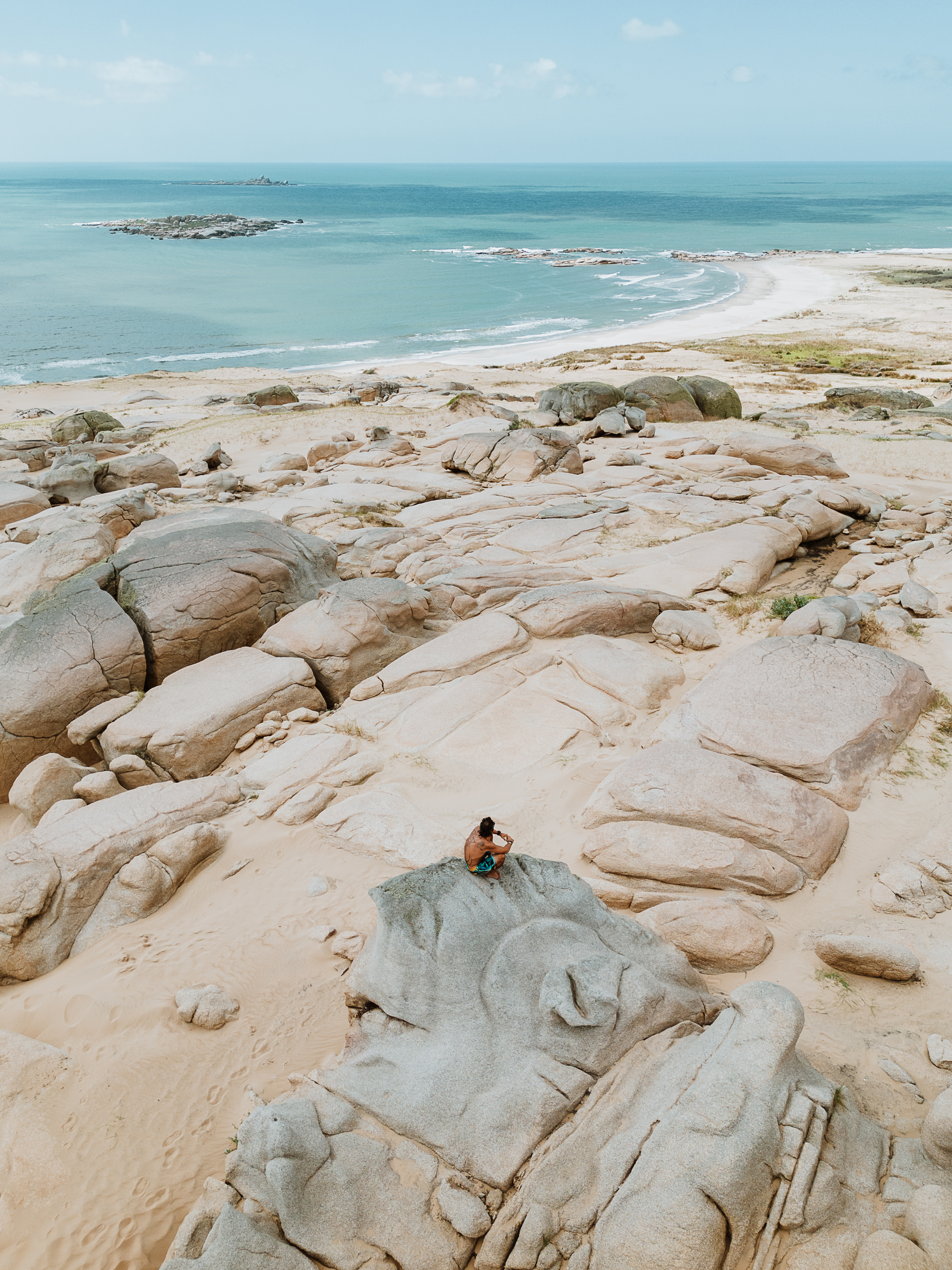 Person sits on pale coastal rocks beside the ocean under a clear sky.