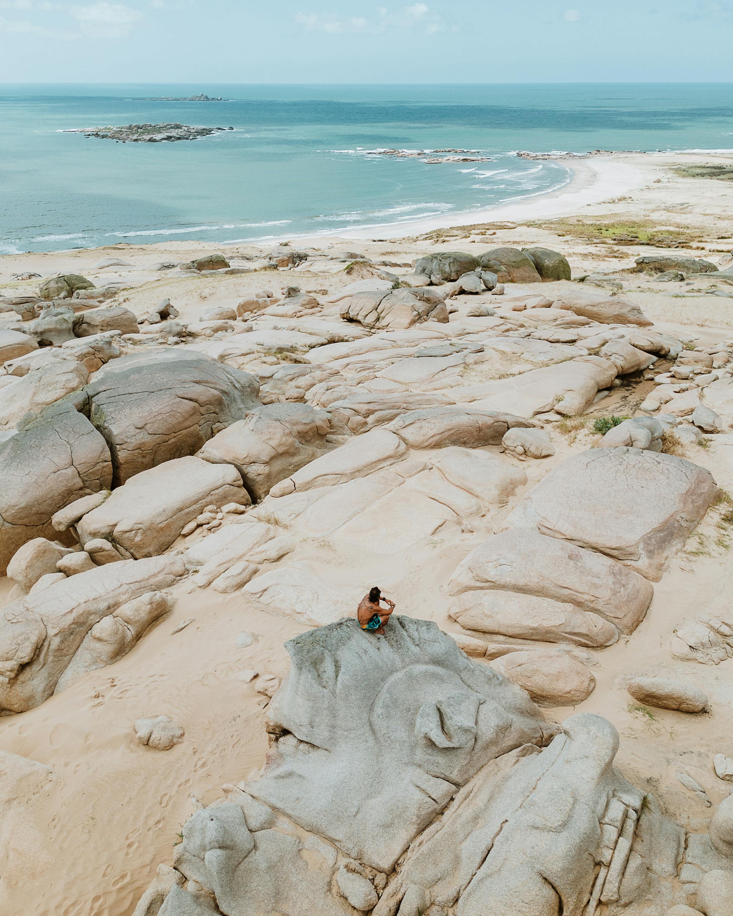 Person sits on pale coastal rocks beside the ocean under a clear sky.