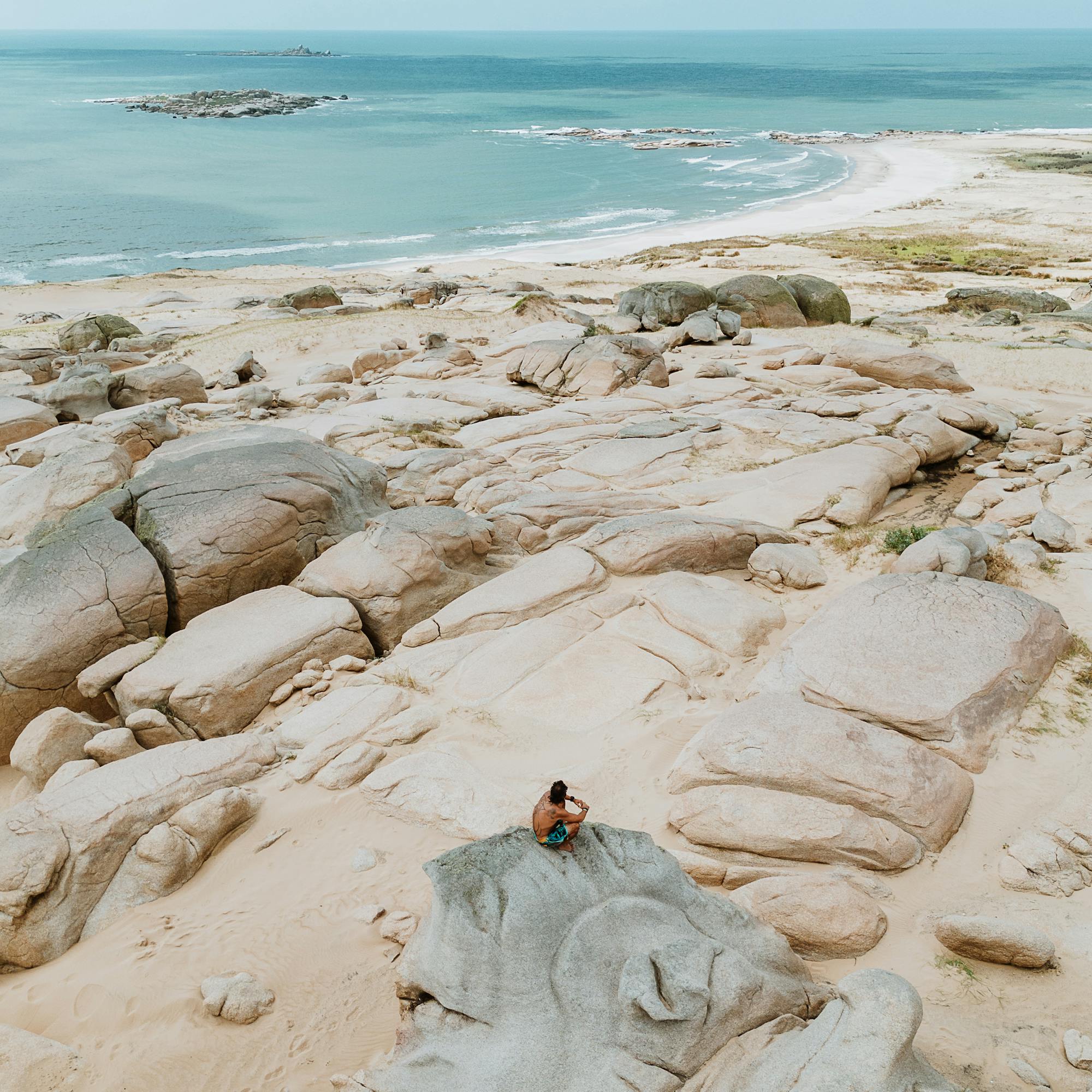 Person sits on pale coastal rocks beside the ocean under a clear sky.