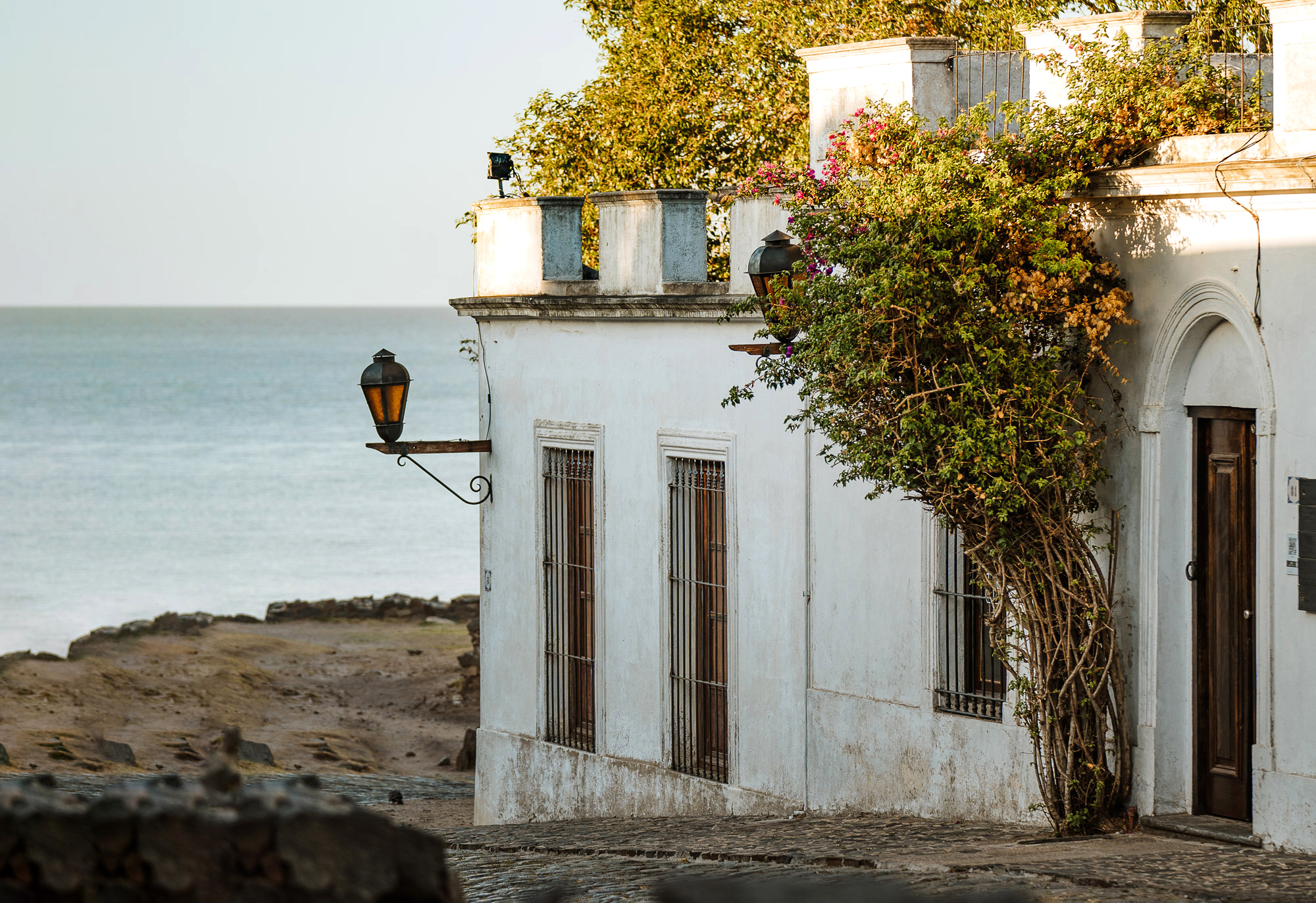 White stucco building with climbing vines stands beside a rocky shoreline with calm water beyond.