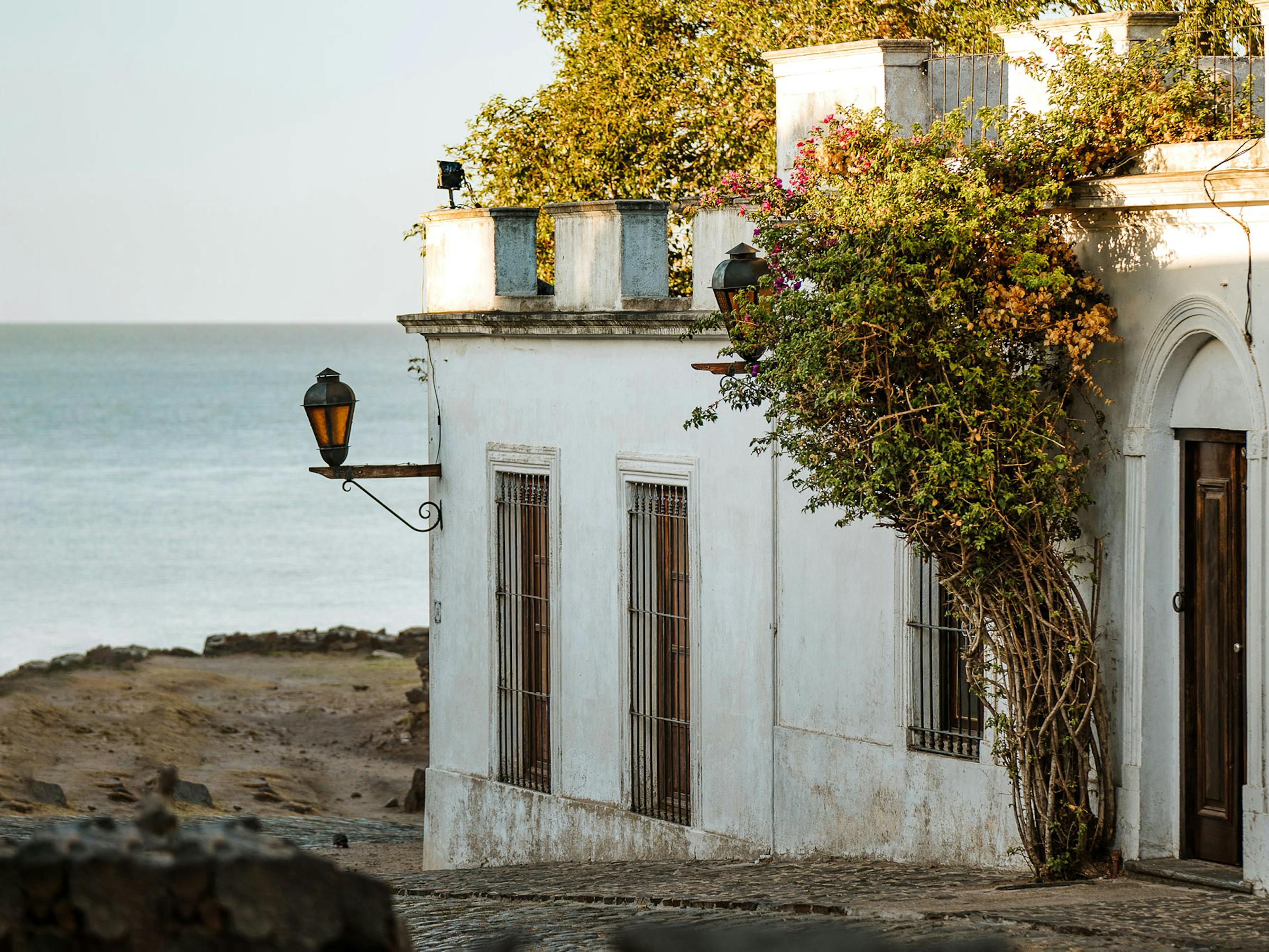White stucco building with climbing vines stands beside a rocky shoreline with calm water beyond.