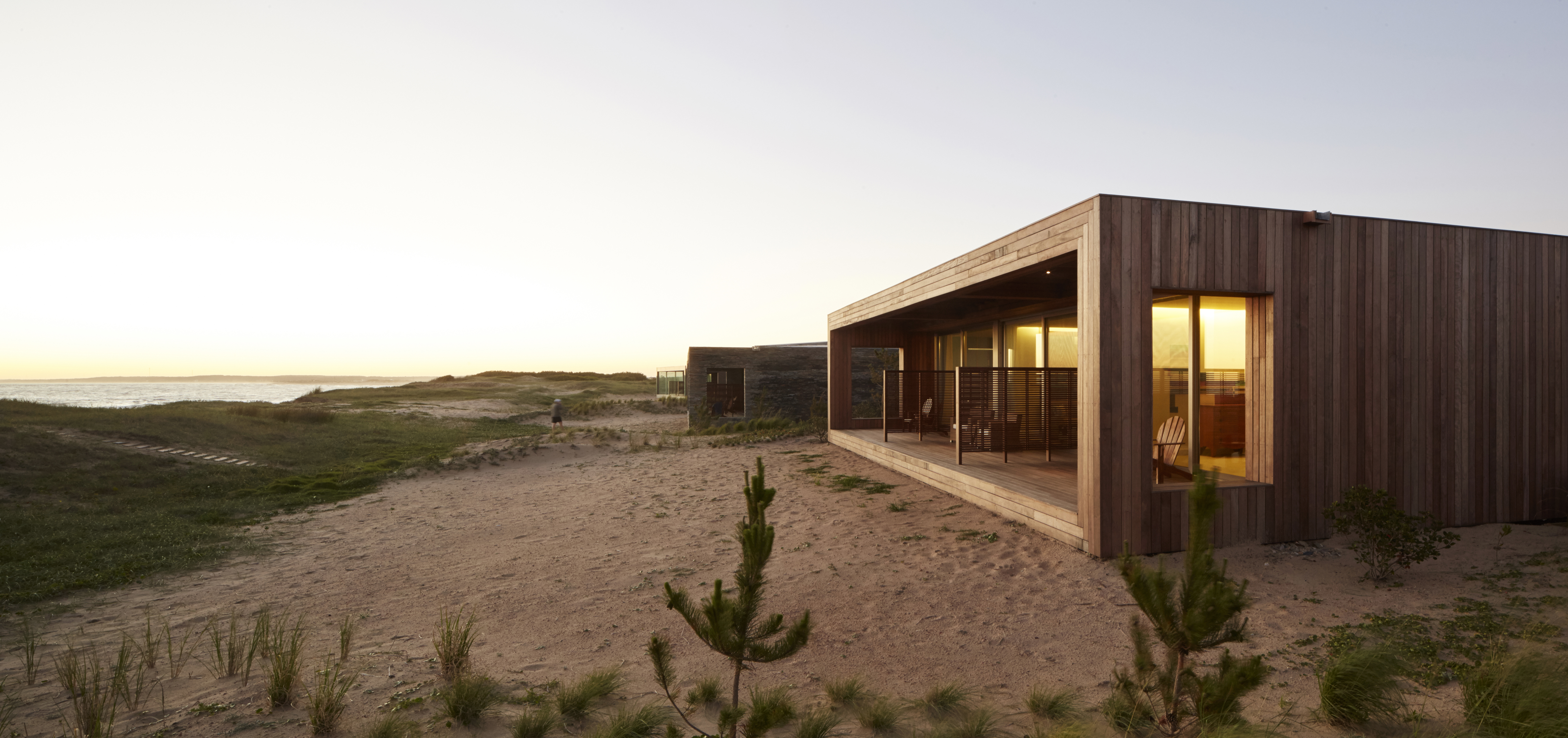 Modern wooden bungalow sits among sandy dunes at sunset with warm light glowing inside.