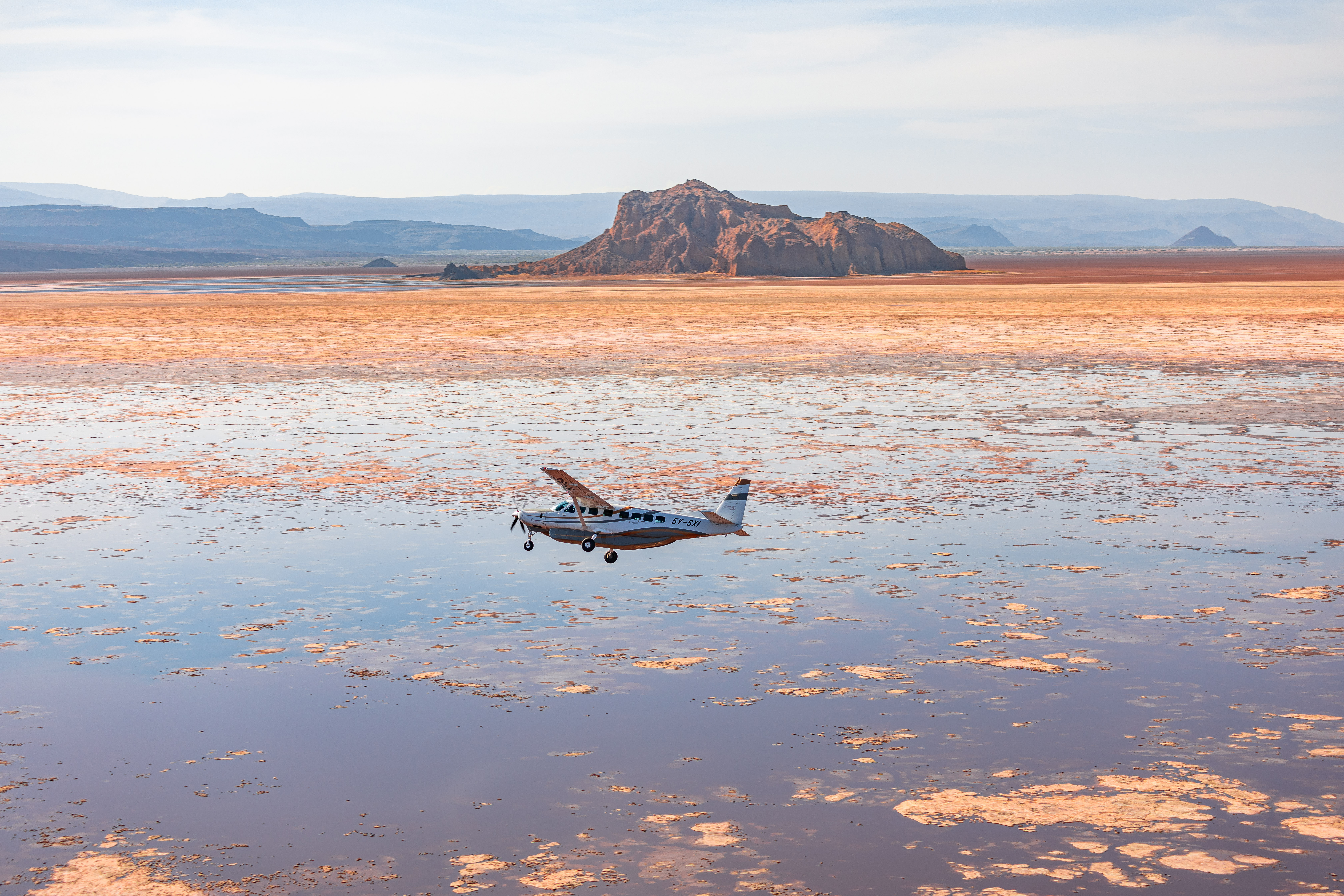 A small plane sits on a reflective salt pan with rippled water, while low mountains rise beneath a pale sky.