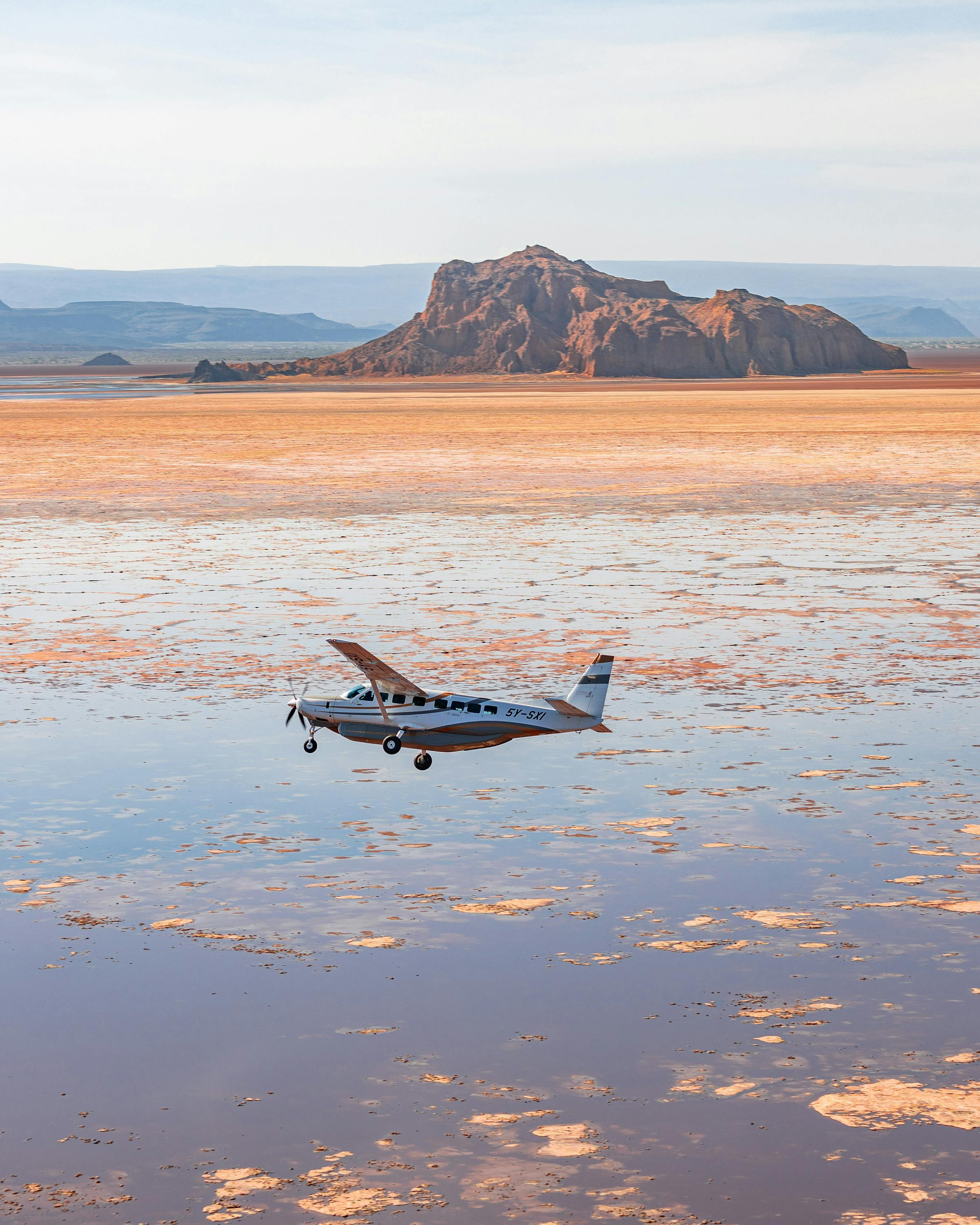 A small plane sits on a reflective salt pan with rippled water, while low mountains rise beneath a pale sky.