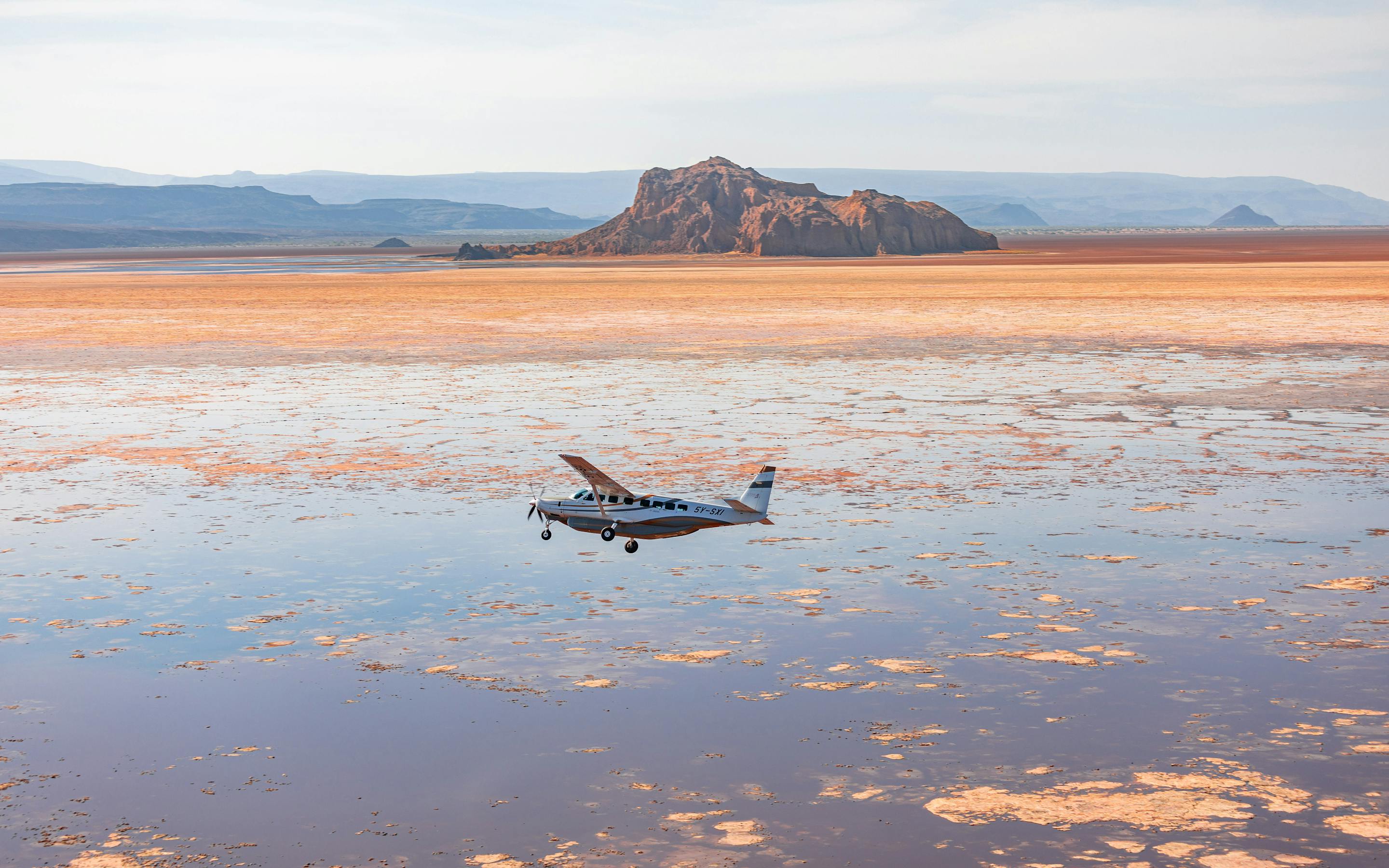 A small plane sits on a reflective salt pan with rippled water, while low mountains rise beneath a pale sky.