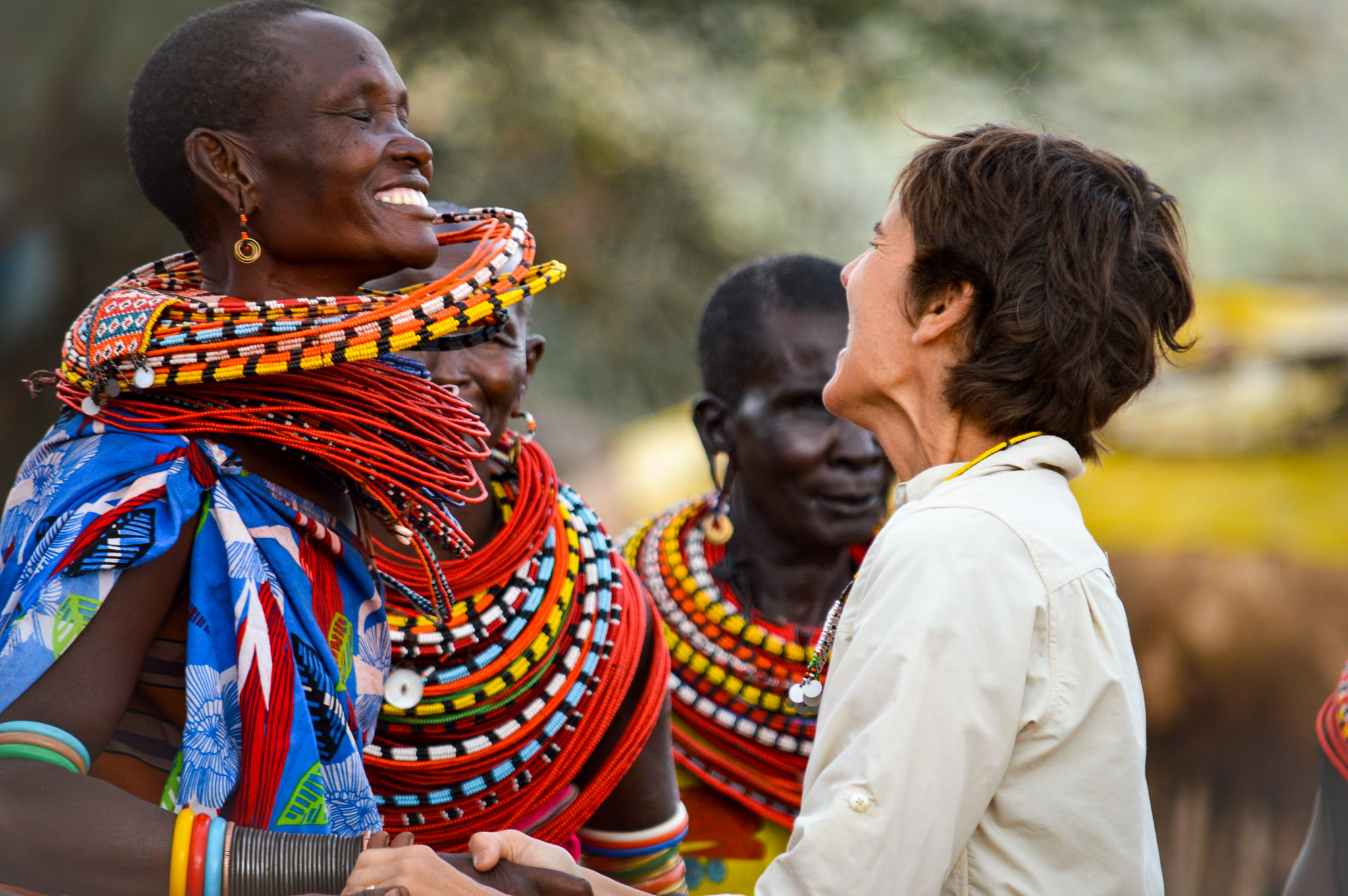 Friends wearing colorful beadwork share smiles while greeting a visitor, framed in a close-up with soft blur.