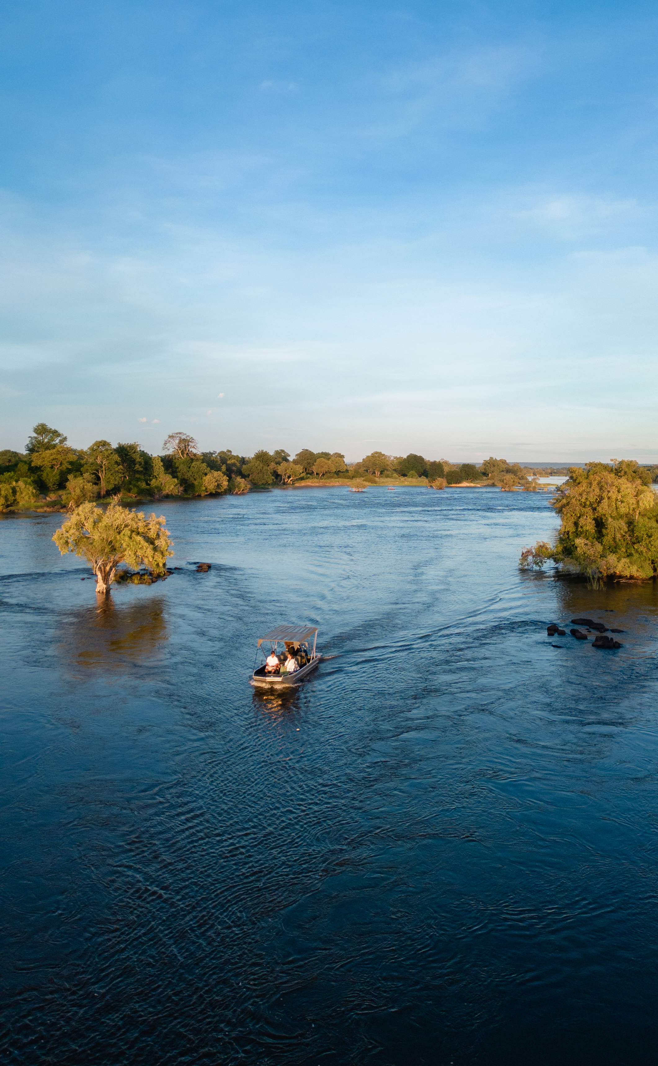 Small motorboats travel along a wide blue river bordered by green trees and low islands beneath a bright, clear sky.