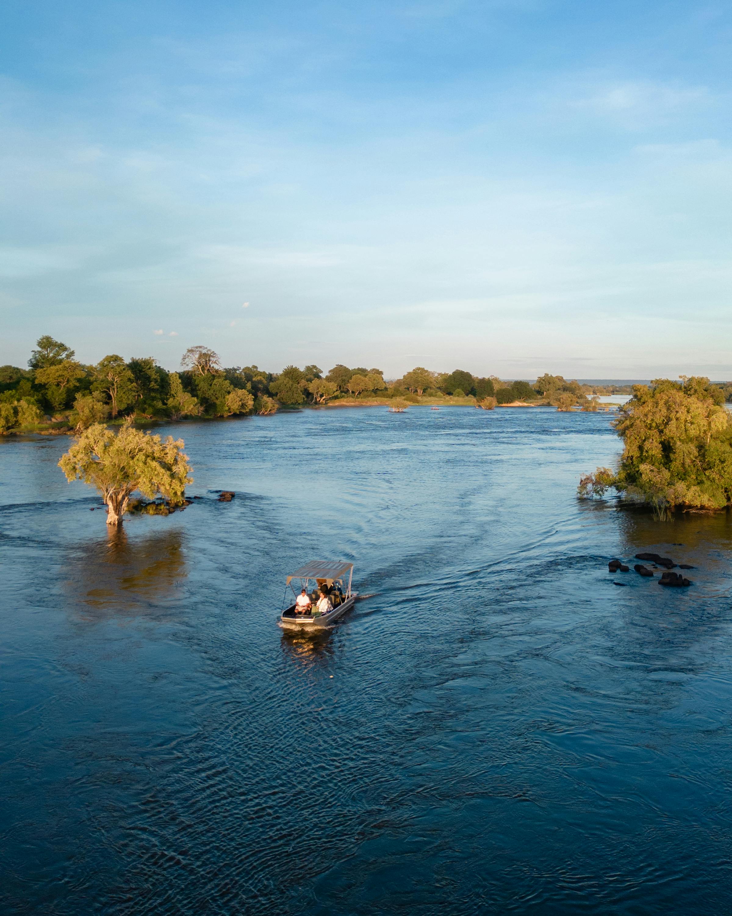 Small motorboats travel along a wide blue river bordered by green trees and low islands beneath a bright, clear sky.