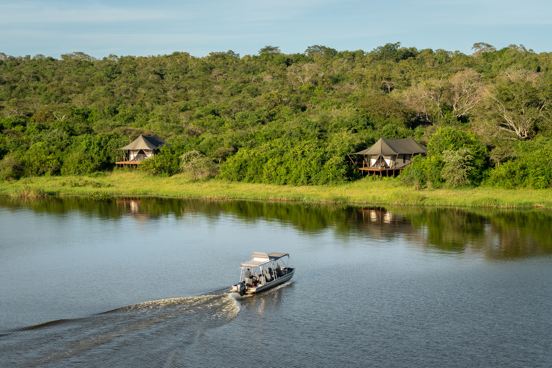 A small motorboat leaves a wake on a calm lake, with shoreline cabins and dense green hills rising behind.