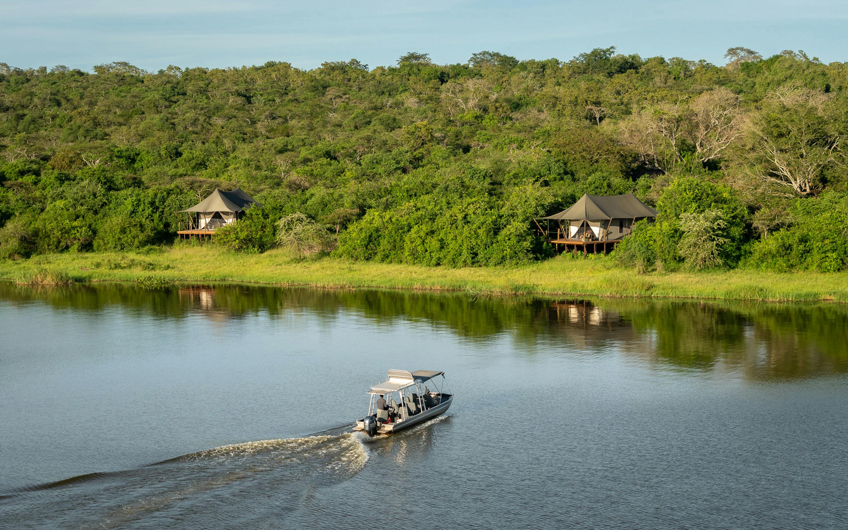 A small motorboat leaves a wake on a calm lake, with shoreline cabins and dense green hills rising behind.