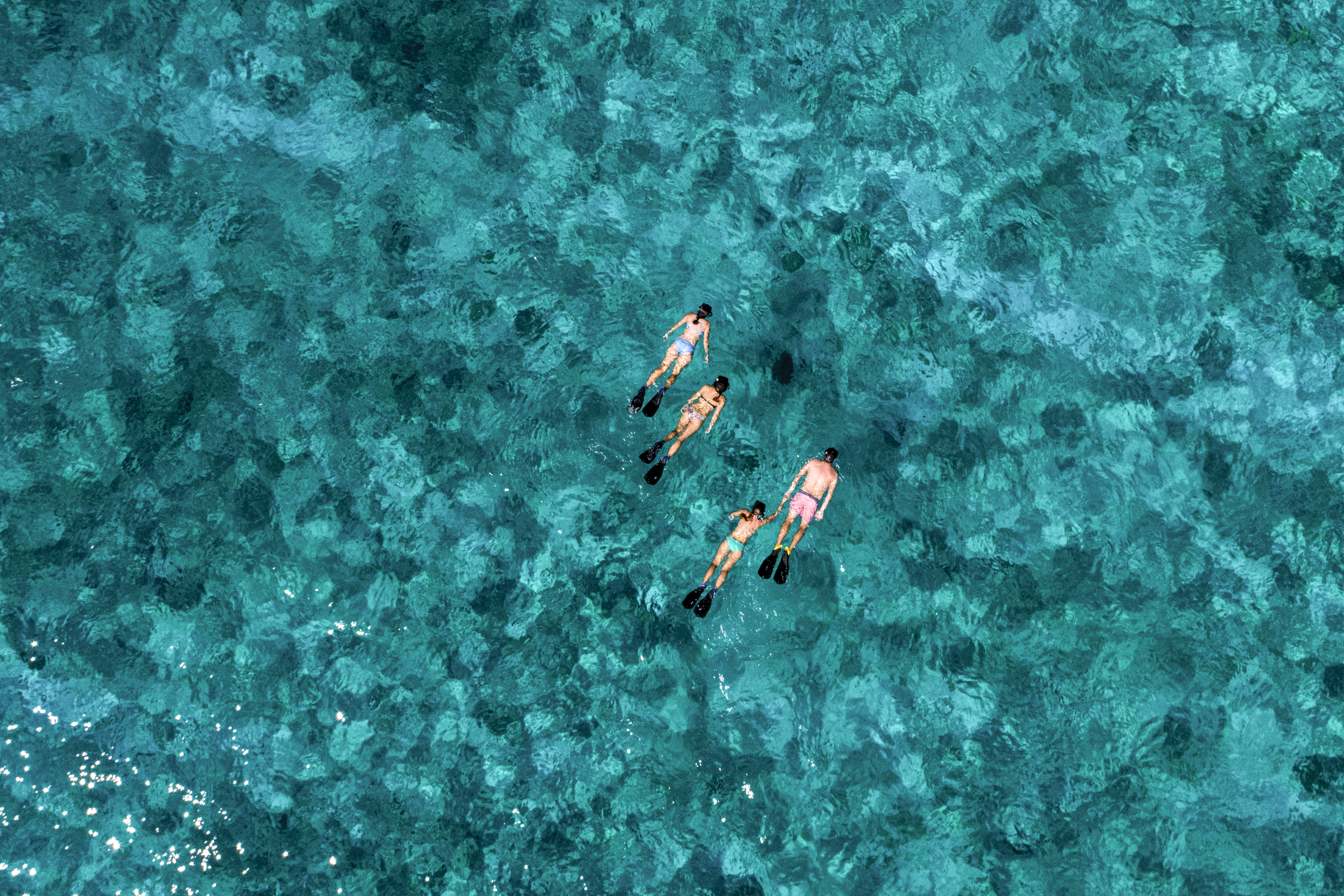 Aerial view of three snorkelers drifting over a turquoise reef, their fins bright against the mottled water.