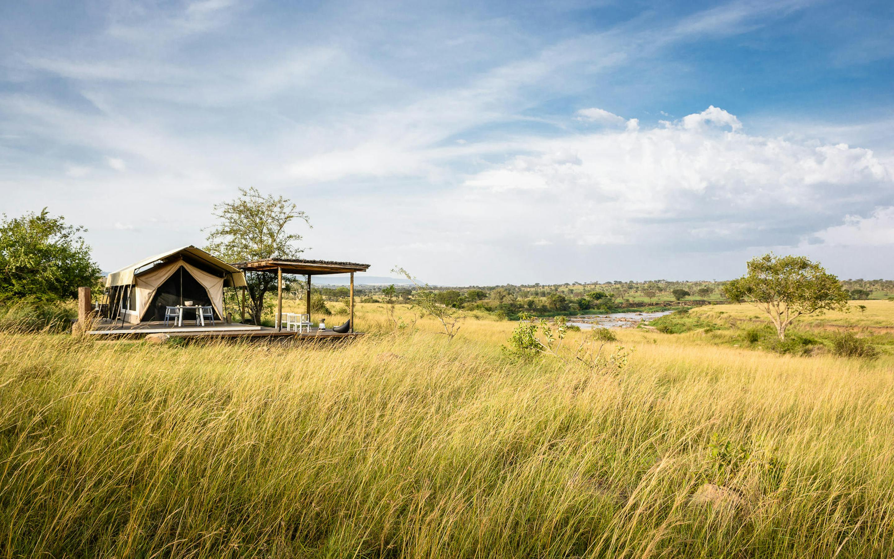 A low tented lodge stands in tall golden grass under a wide sky, with open plains and scattered trees beyond.