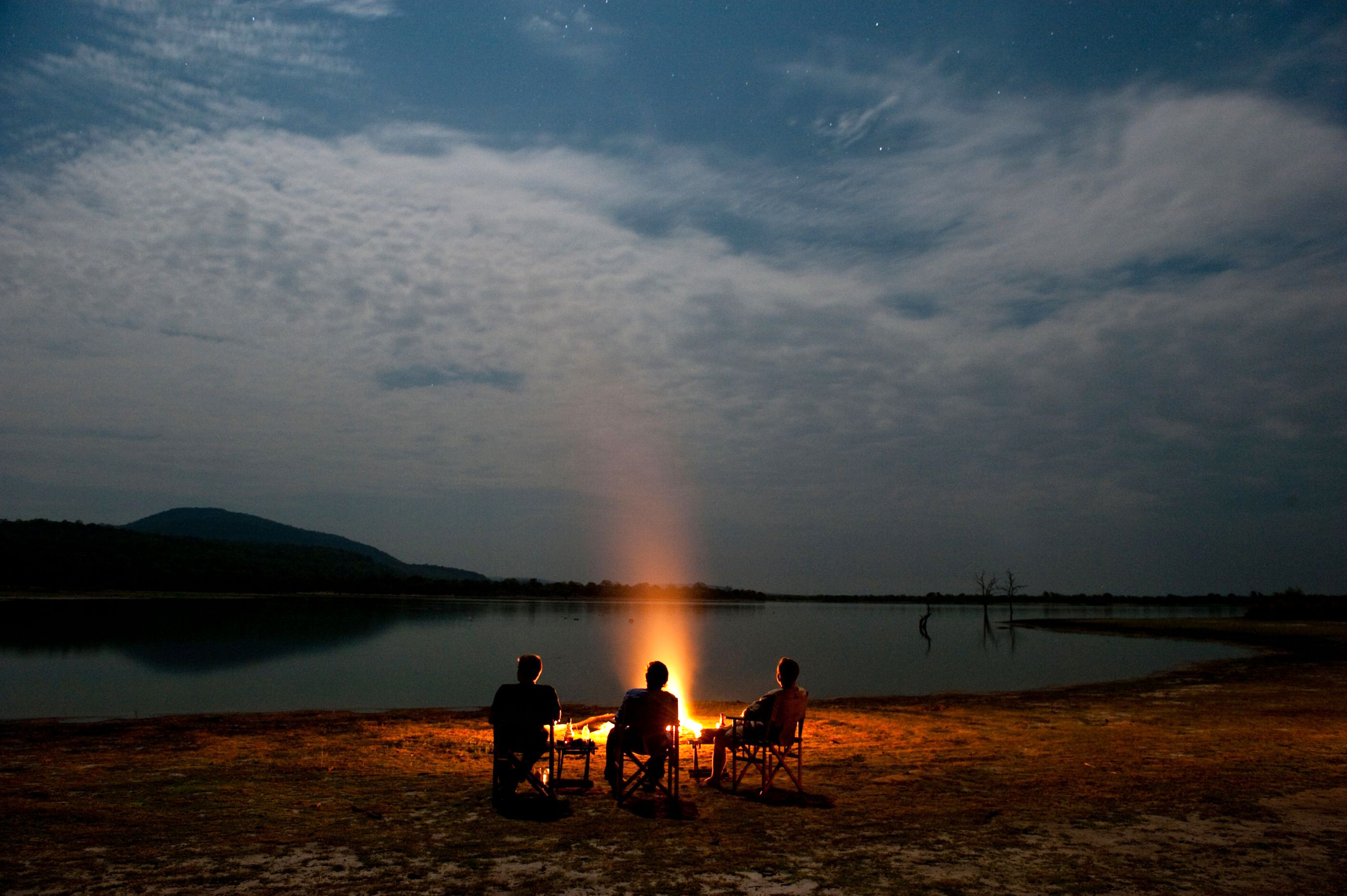 Silhouetted people sit around a campfire on a sandy lakeshore at dusk beneath dramatic clouds and a faint horizon glow.