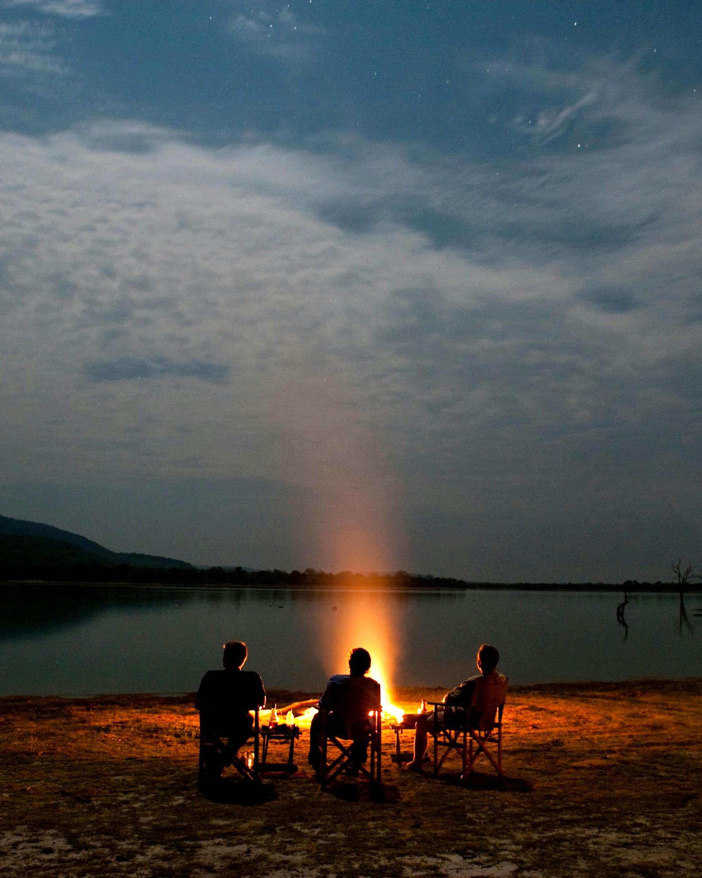 Silhouetted people sit around a campfire on a sandy lakeshore at dusk beneath dramatic clouds and a faint horizon glow.