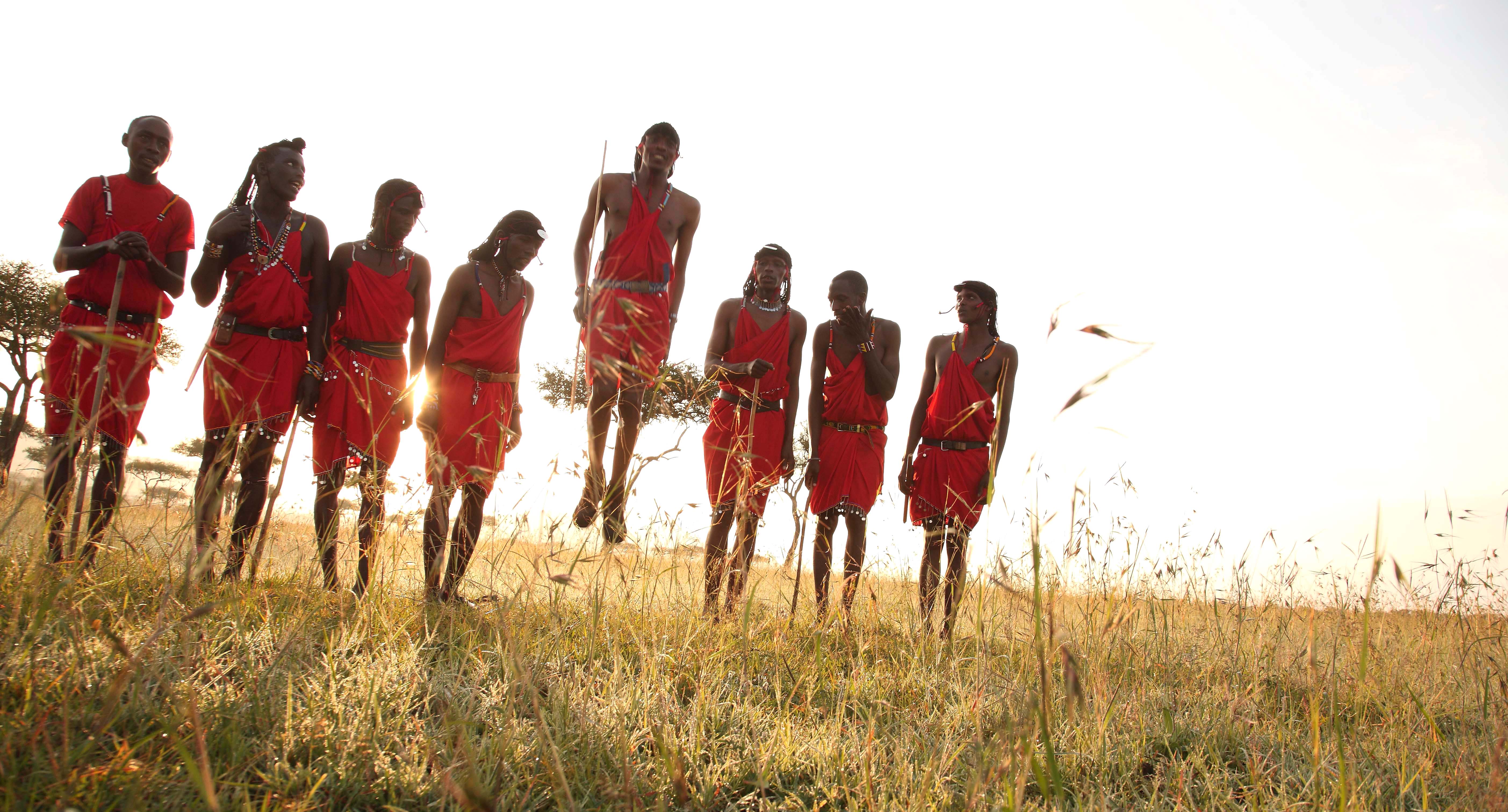 A line of people in red robes jump on a grassy ridge at sunset, backlit by warm light against a bright sky.