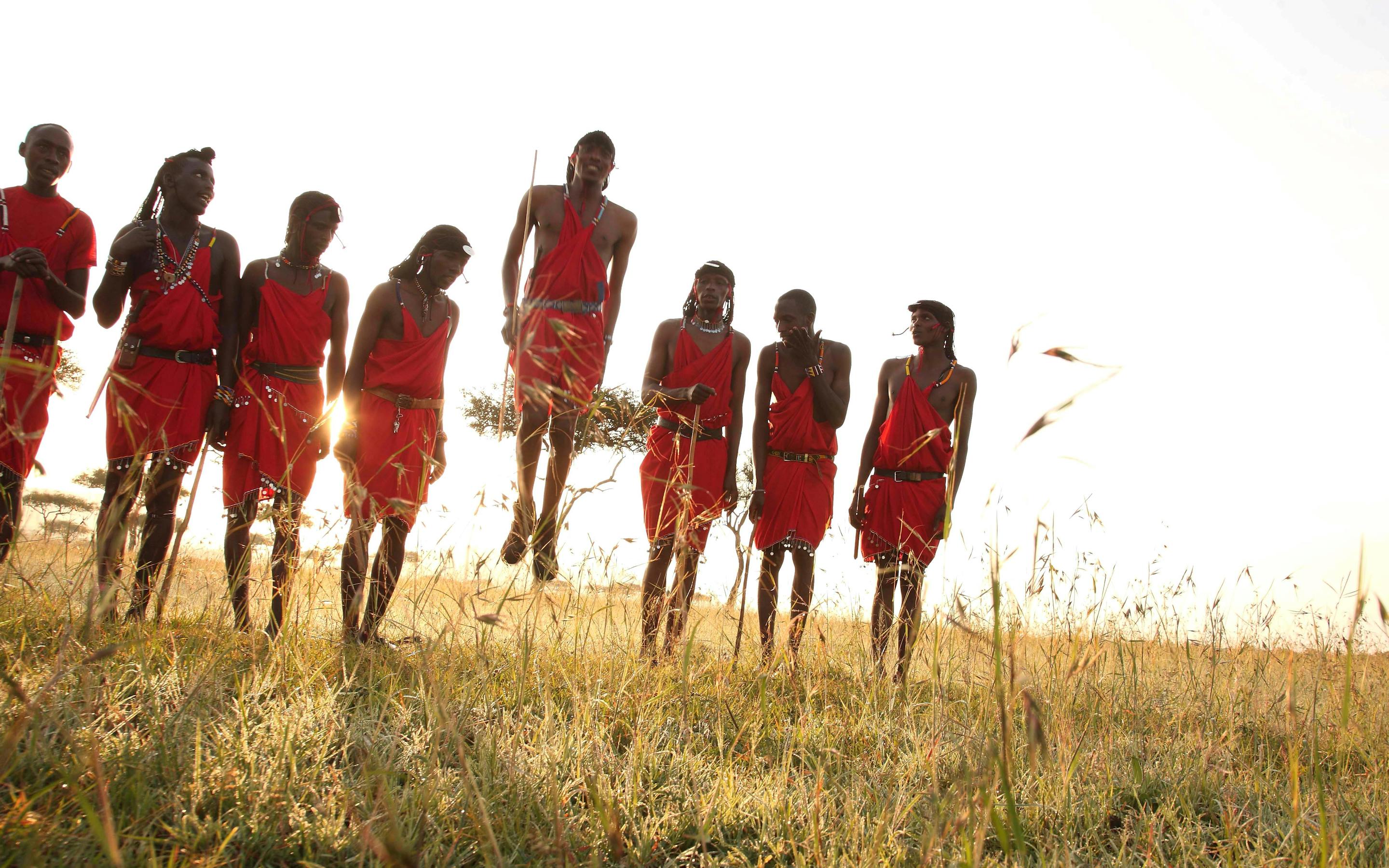 A line of people in red robes jump on a grassy ridge at sunset, backlit by warm light against a bright sky.