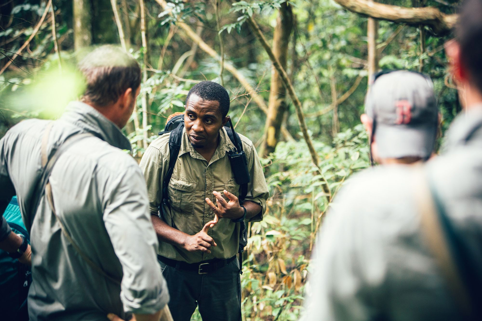 A small group talks on a narrow forest trail, one person gesturing while others listen among green leaves.