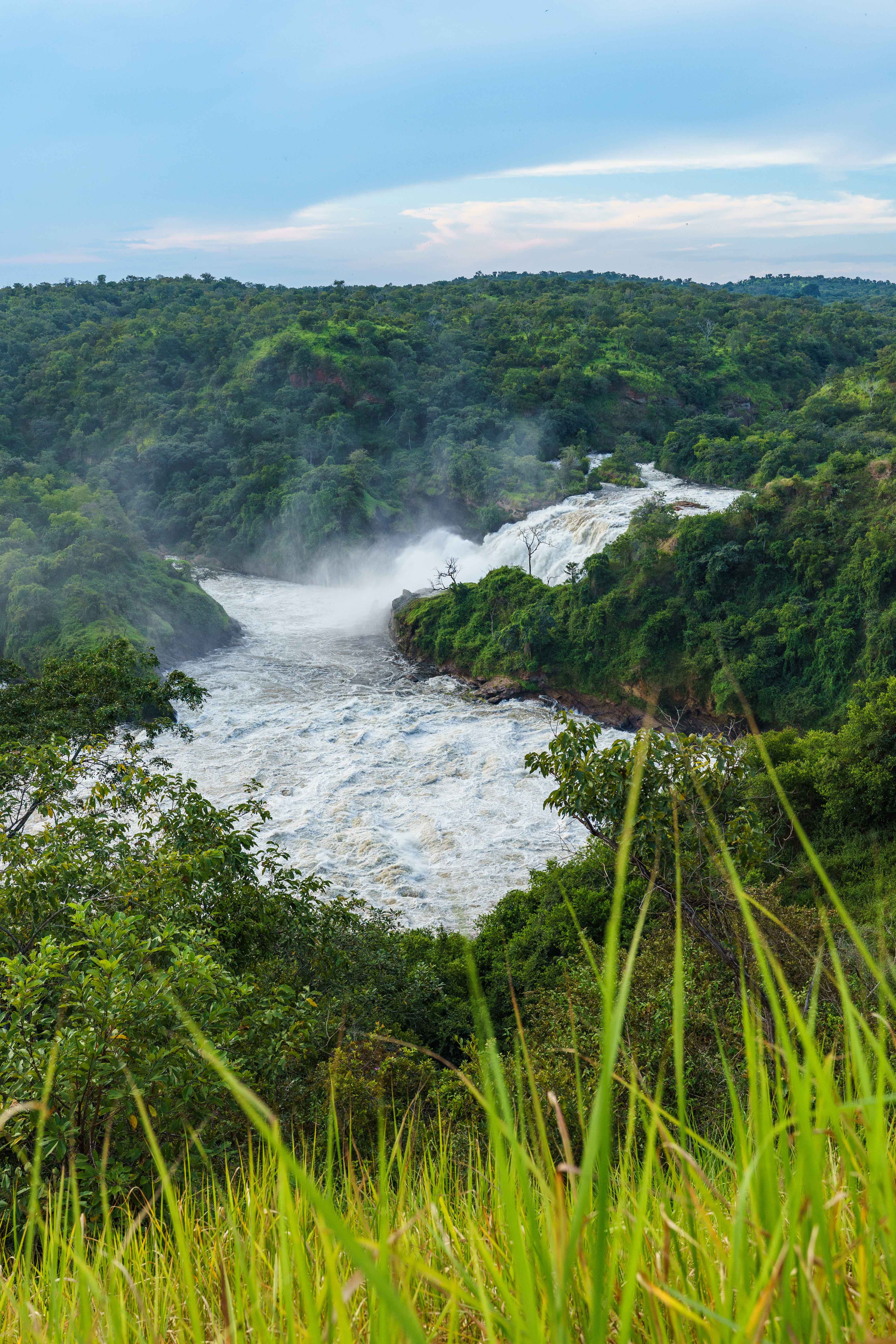 A wide river churns through a misty valley of rolling green hills, with white spray rising above the rapids.