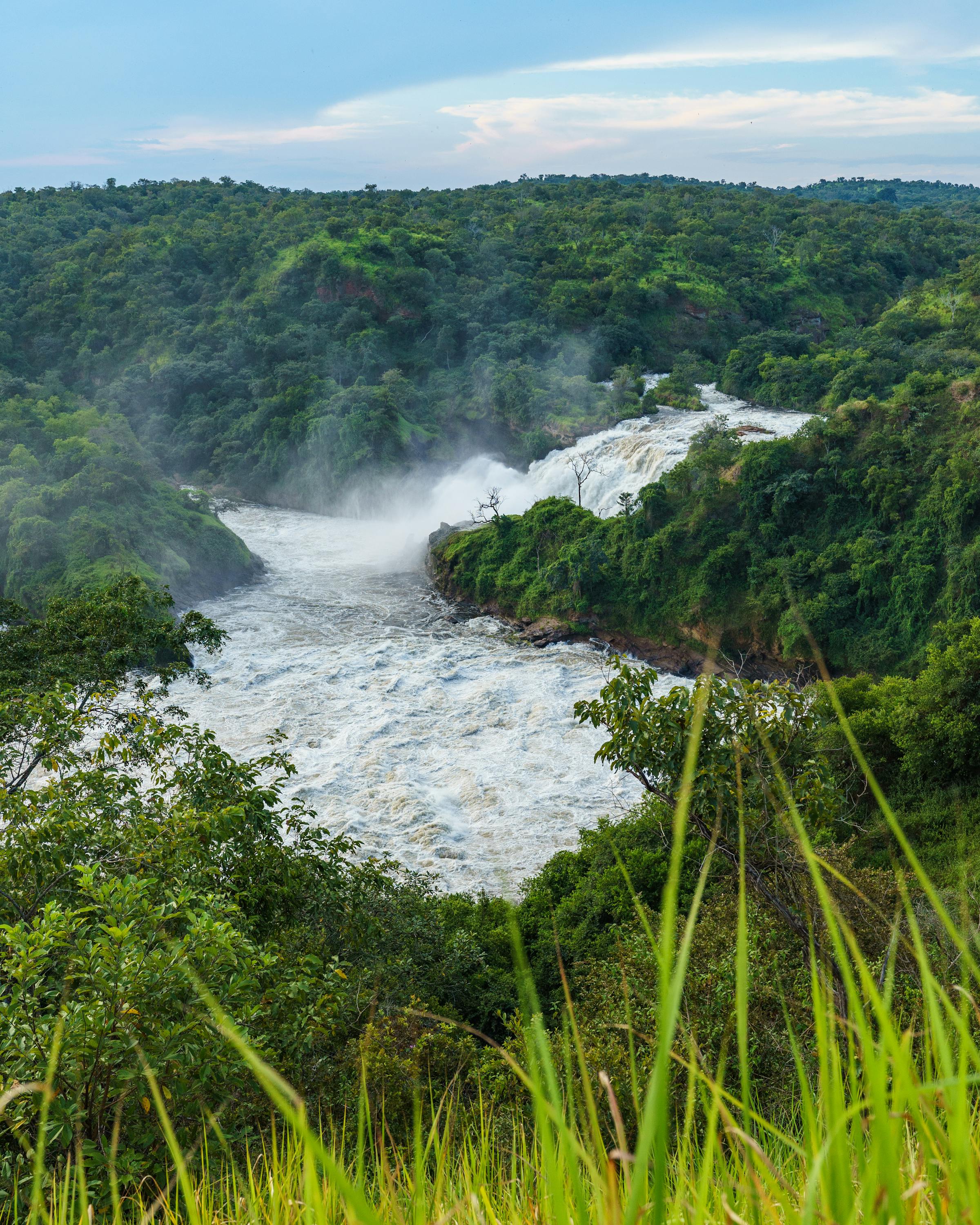 A wide river churns through a misty valley of rolling green hills, with white spray rising above the rapids.